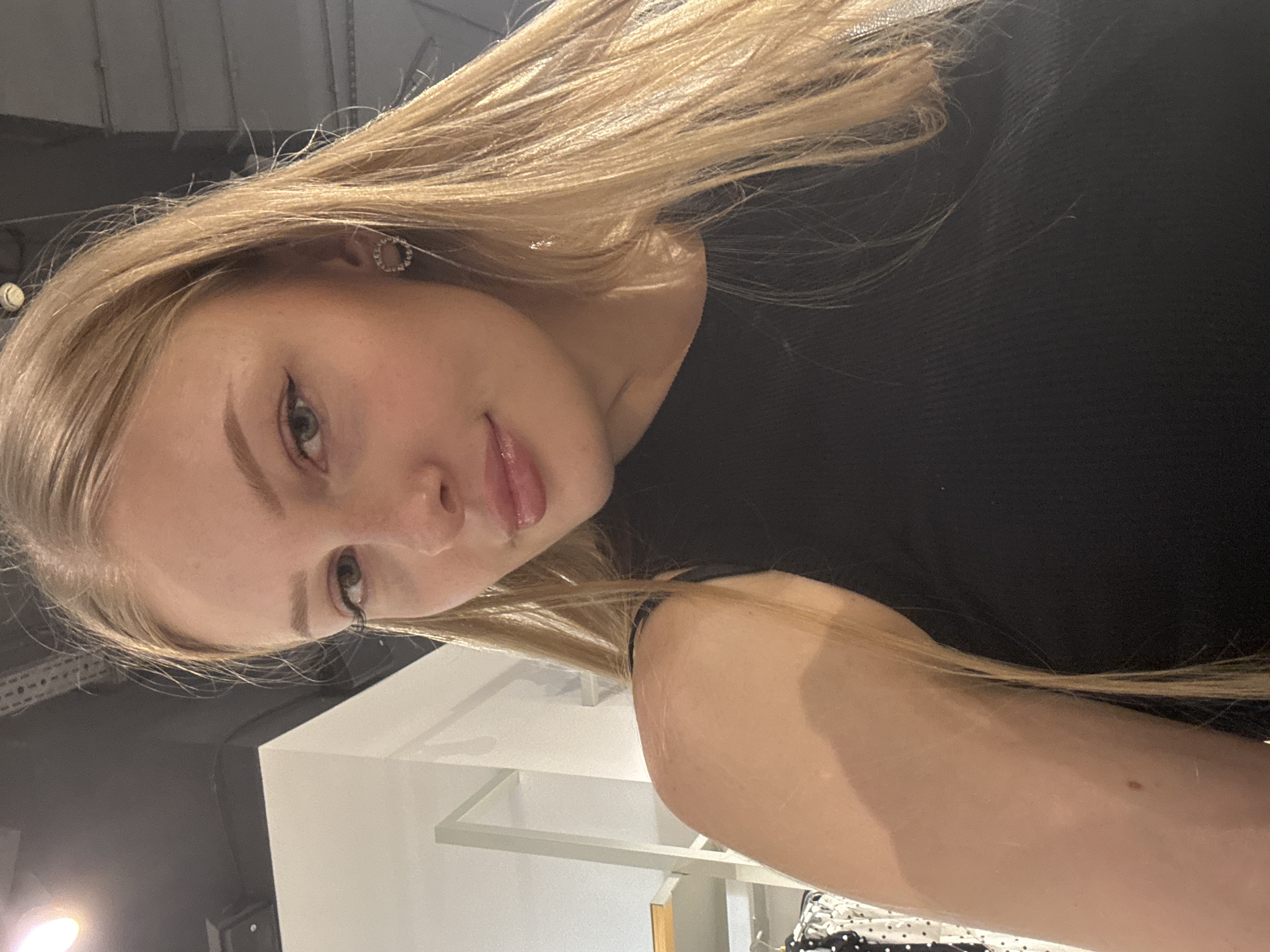 A young woman with blonde hair and blue eyes is captured in a close-up, selfie-style photo, with her face angled slightly to the right. She is wearing a black, sleeveless top and a small, circular earring adorned with rhinestones in her left ear. Her lips are slightly parted, and she is wearing subtle makeup, including eyeliner and lip gloss. The background suggests an indoor retail environment, possibly a clothing store, with white shelving units and racks of clothing visible to the left, and industrial ceiling elements in the upper portion of the frame. The lighting is warm, casting a soft glow on her skin and hair. There is no indication of the city of Perm, Russia, in the image.