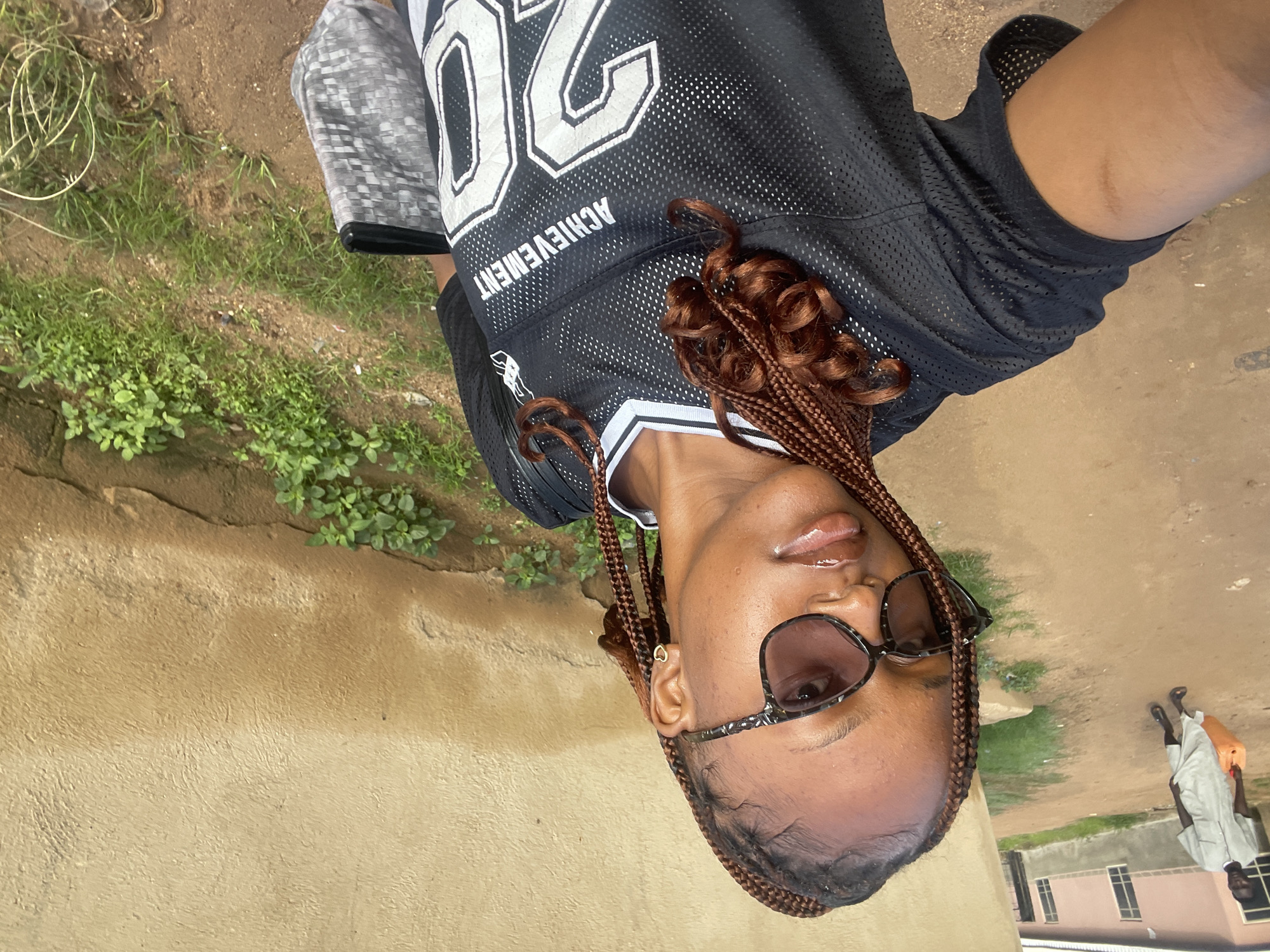 A young woman with braided hair and sunglasses is taking a selfie outdoors. She is wearing a black athletic jersey with white lettering and trim, and she is holding a patterned bag. Behind her, a man in a light-colored robe carries a large orange container. The background shows a dirt path, a building with windows, and a textured wall with green vegetation growing at its base. This scene appears to be located in Jos, Nigeria.
