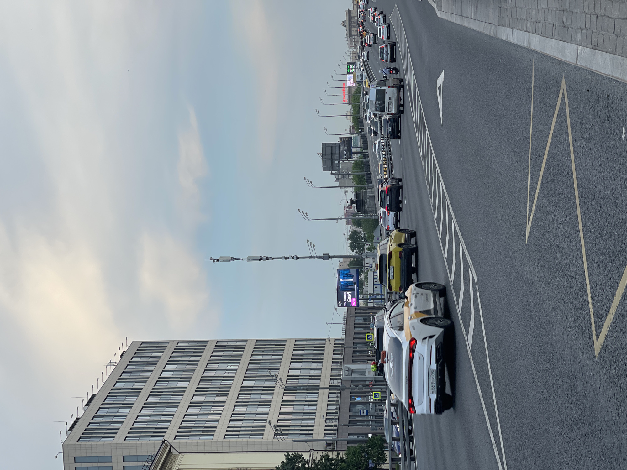 The image depicts a congested urban roadway in Unknown, Unknown. The road is divided into multiple lanes, with heavy traffic consisting mainly of cars and vans. On the left side of the road stands a modern building with numerous windows. Streetlights and billboards are visible along the road, and the sky is overcast.