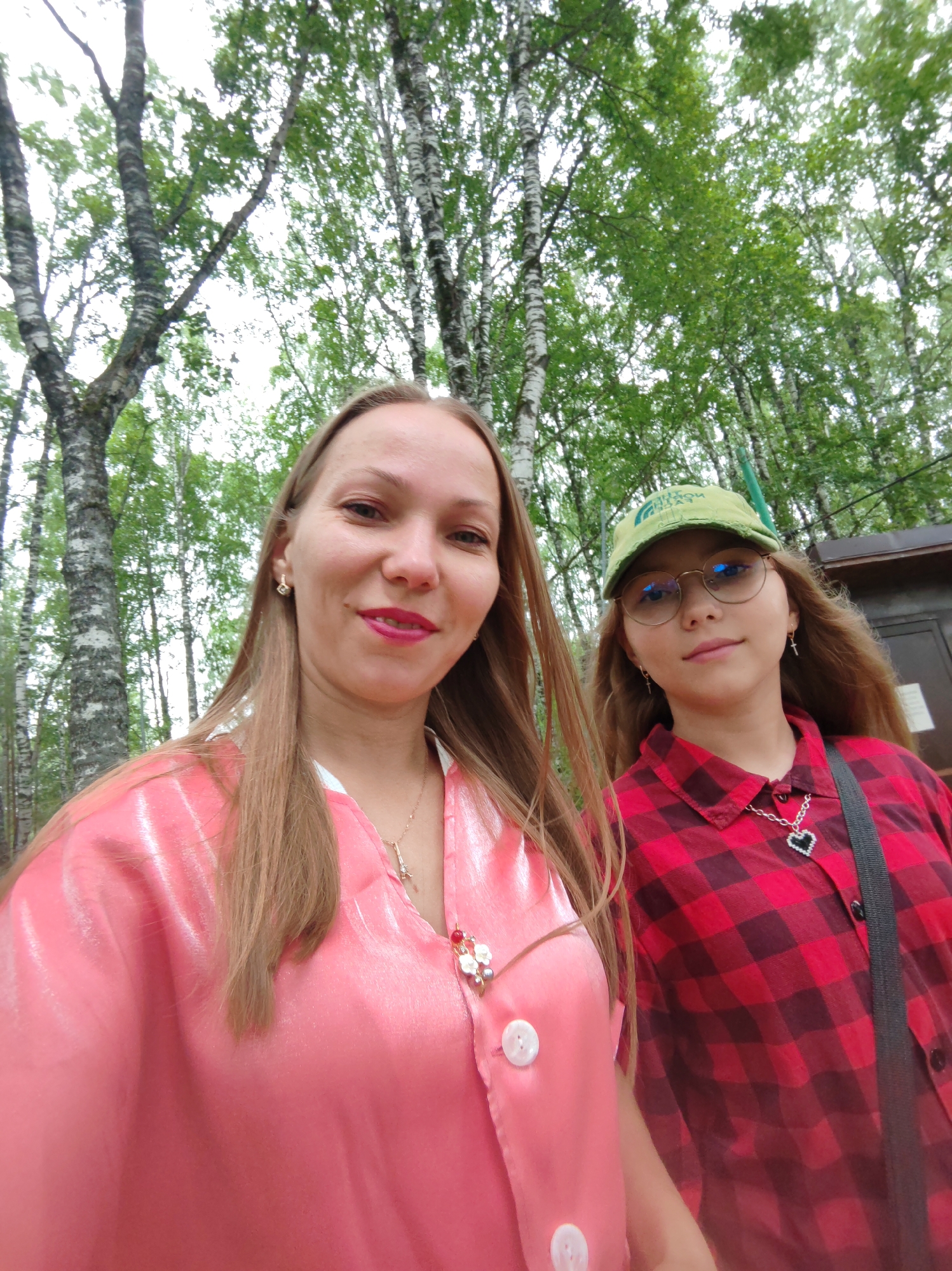 Two individuals, a woman and a young girl, are posing for a selfie in a park setting. The woman, on the left, has long blonde hair, is wearing a shiny pink jacket with white trim and buttons, and a necklace. She is smiling and looking at the camera. The young girl, on the right, is wearing a green baseball cap, glasses, and a red and black plaid shirt. She also has a necklace with a heart pendant. They are standing in front of a background of birch trees with green foliage, suggesting a summer or spring day. The image appears to have been taken outdoors in a natural environment, possibly a park in Syktyvkar, Russia, given the prevalence of birch trees in the region and the overall aesthetic.