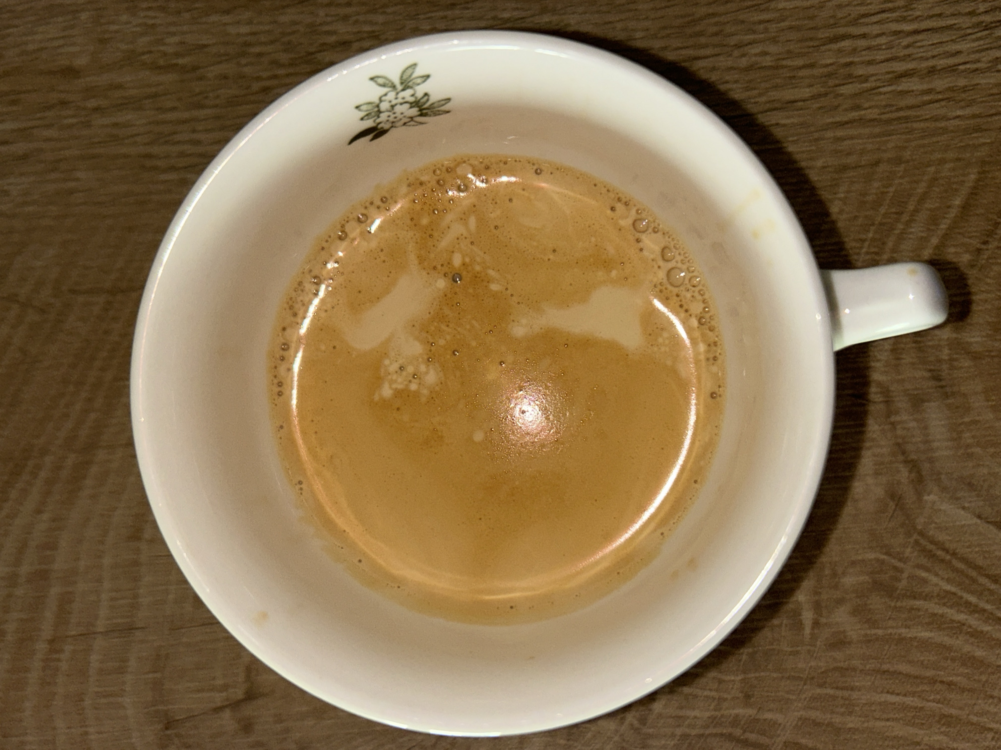 A close-up, overhead shot shows a white porcelain cup filled with coffee, likely an espresso or latte. The cup features a small floral design near the top edge on the inside. The coffee has a light brown color with foamy bubbles around the edges. The cup has a small handle on the right. The cup sits on a wooden surface with a grainy texture.
There is no visual information to suggest that the photo was taken in Plovdiv, Bulgaria, or any other specific location.