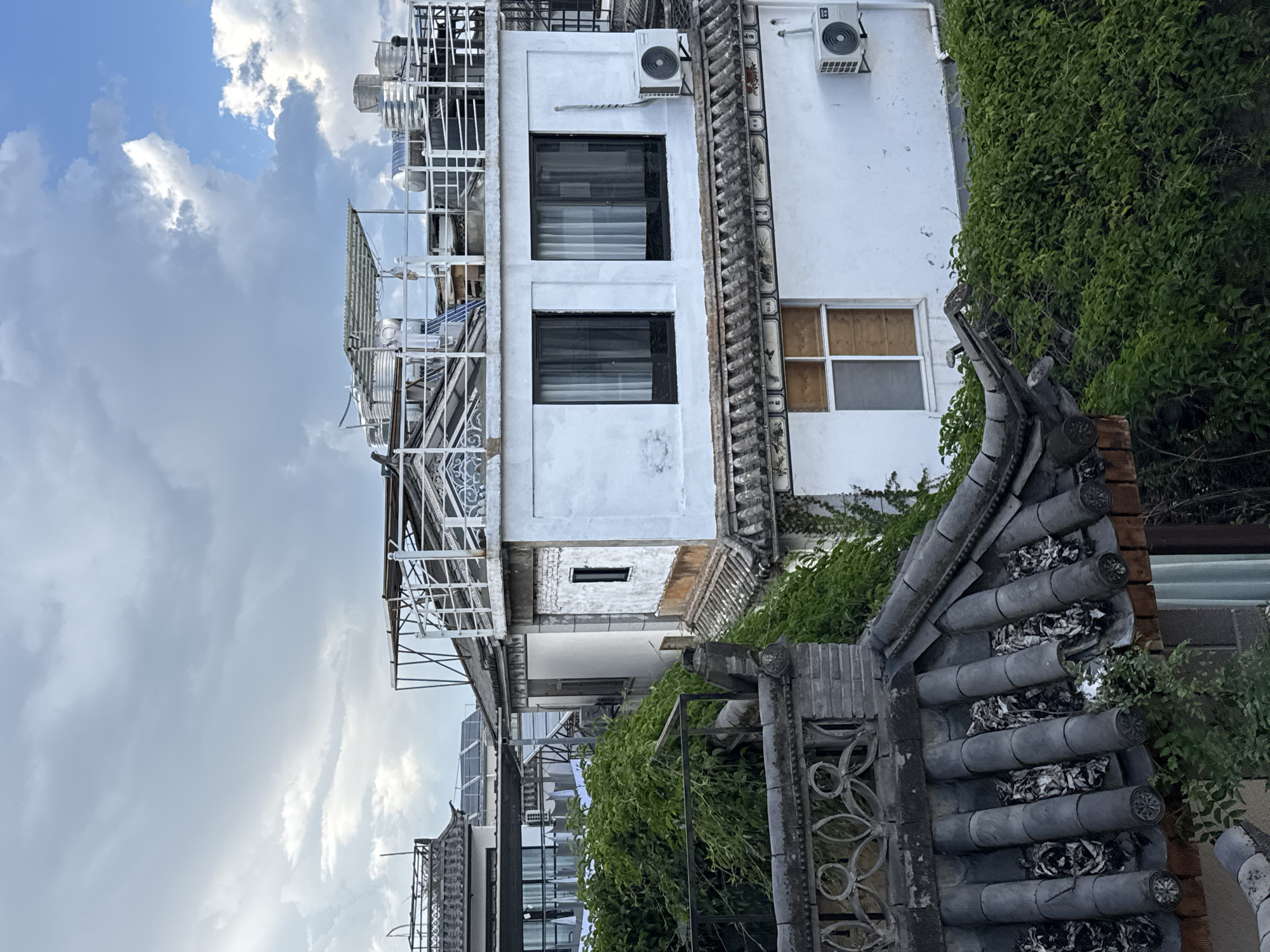This is a medium-range, eye-level shot of rooftops in Dali City, China, under a cloudy sky. The scene is dominated by traditional Chinese tiled roofs and white stucco buildings. Lush green foliage cascades over some of the structures, creating a sense of overgrown nature reclaiming the urban landscape.
In the foreground, a dark tiled roof with distinctive curved eaves and ornate brickwork is visible. The tiles themselves appear weathered and somewhat damaged in places, with dried leaves or debris lodged between them. To the right, a dense wall of vibrant green leaves obscures much of the building behind it.
Further back, a white, multi-story building with a flat rooftop stands out. This building features two large, dark-framed windows on its upper level, with sheer white curtains drawn inside. An air conditioning unit is mounted on the wall below the windows. Another, smaller window with a wooden frame and frosted glass is located on the lower level of this building. A second, smaller air conditioning unit is visible on the lower part of this wall.
The rooftop of the white building is complex, with a white metal railing surrounding the perimeter. Several large, silver water tanks are present on the rooftop, along with a corrugated metal canopy supported by metal poles. A partially visible structure with a more industrial, modern aesthetic is situated to the far right, behind the white building.
In the background, more rooftops and buildings are visible, including some with dark tiled roofs and what appear to be solar panels on one of them. The sky above is a mix of grey clouds and patches of bright blue, suggesting the weather is variable. There are no people visible in the image. The overall impression is one of a residential or commercial area in an older part of Dali City, where traditional architecture is interwoven with elements of modern infrastructure and nature.