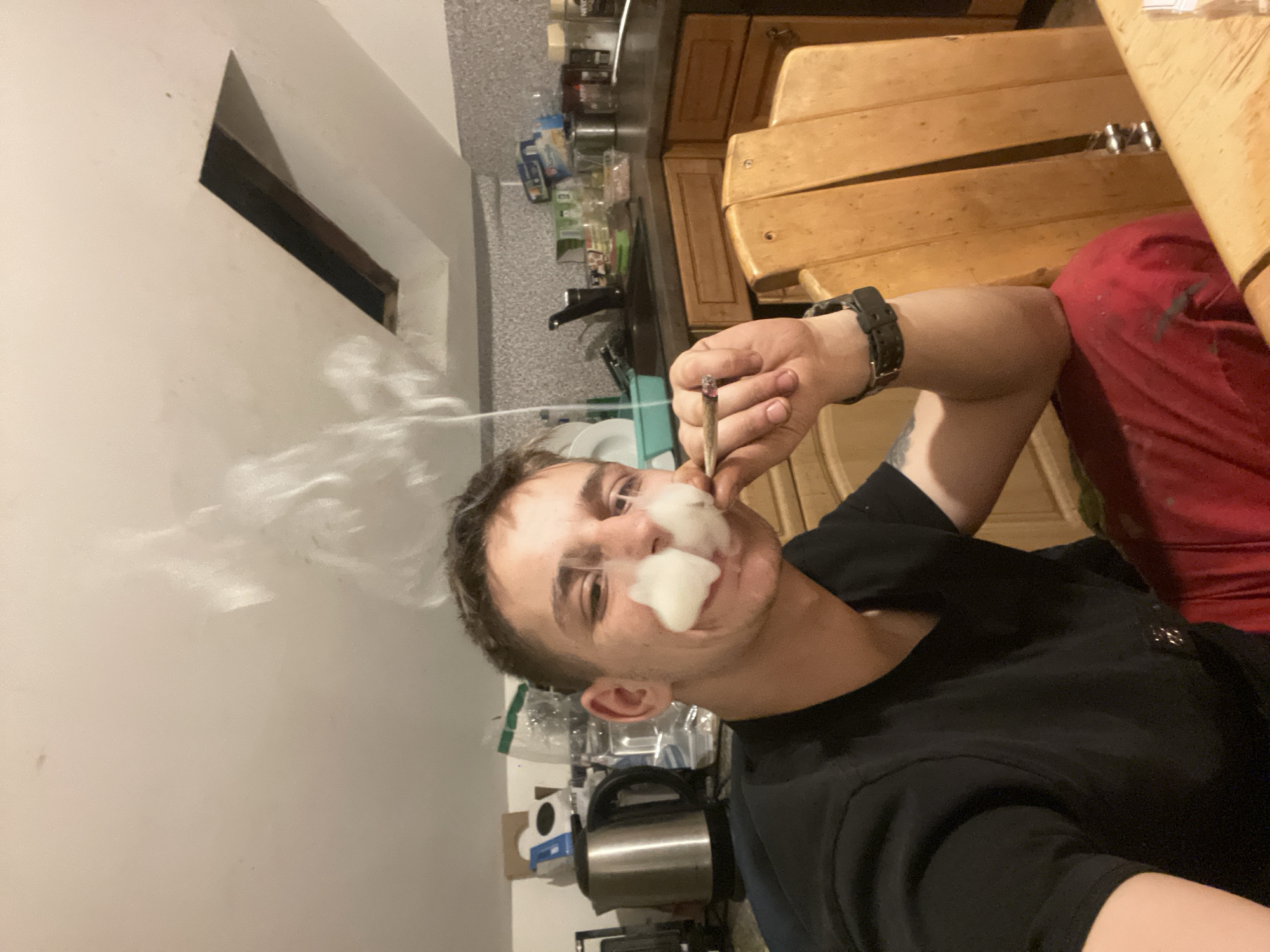 This image captures a close-up indoor scene within a kitchen in Unknown, Unknown. A male individual, appearing to be in his 20s to 30s, is centered in the frame, taking a selfie. He has short, dark, textured hair and is wearing a black short-sleeved t-shirt and red pants. A dark-colored watch is visible on his right wrist, and a faint tattoo is present on his left forearm. He is smiling faintly and holding a lit, hand-rolled cigarette or joint between his right thumb and forefinger near his mouth. A distinct plume of white smoke is exhaled from his mouth and nostrils, forming two discernible cloud-like shapes near his nose before dissipating upwards.
The background reveals a kitchen area with light-toned wooden base cabinets and a dark, possibly speckled, countertop. The backsplash features a textured, light gray patterned wallpaper or tile. Various kitchen items are visible on the counter, including a silver electric kettle, a large clear plastic water bottle, a white plastic dispenser, and several smaller containers, some metal, some plastic. A light blue plastic basin containing dishes is also present. Food packaging, including a "Nescafe" jar, can be seen on the right side of the counter.
The ceiling above is light-colored and angled, indicating an attic or sloped roof structure. A rectangular cutout in the ceiling, appearing as a dark void, is present on the right side. A portion of a light-colored wooden chair and table is visible in the bottom right corner of the image. The ambient lighting appears to be artificial.