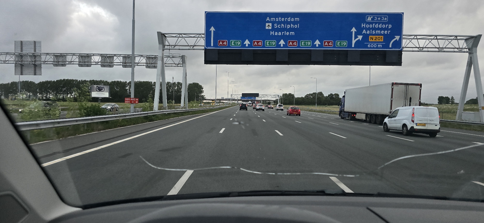 The image provides a driver's perspective from within a vehicle traveling on a multi-lane highway in Hoofddorp, Netherlands, under an overcast sky. The bottom portion of the frame shows the car's dashboard and a cracked windshield.
Ahead, the highway features several vehicles, including a red hatchback, a silver sedan, other cars, a white cargo van with license plate V-93-GJG, and a blue semi-trailer truck with a white trailer.
A prominent blue overhead gantry sign spans the highway, directing traffic. The main part of the sign indicates the route straight ahead for "Amsterdam," "Schiphol," and "Haarlem" via motorways A4 and E19. To the right, an exit sign for "Hoofddorp" and "Aalsmeer" is displayed, designated as exit "3 + 3a" and national road "N 201," with a notice indicating the exit is "600 m" away.
Along the left side of the road, behind a metal guardrail and a line of green trees, are smaller red and white roadside signs reading "UGS ABD ↑" and "UGS C →." Further in the distance, a billboard is visible, depicting what appears to be a car wheel or steering wheel. Streetlights line the highway, and the overall weather appears to be cloudy daytime.