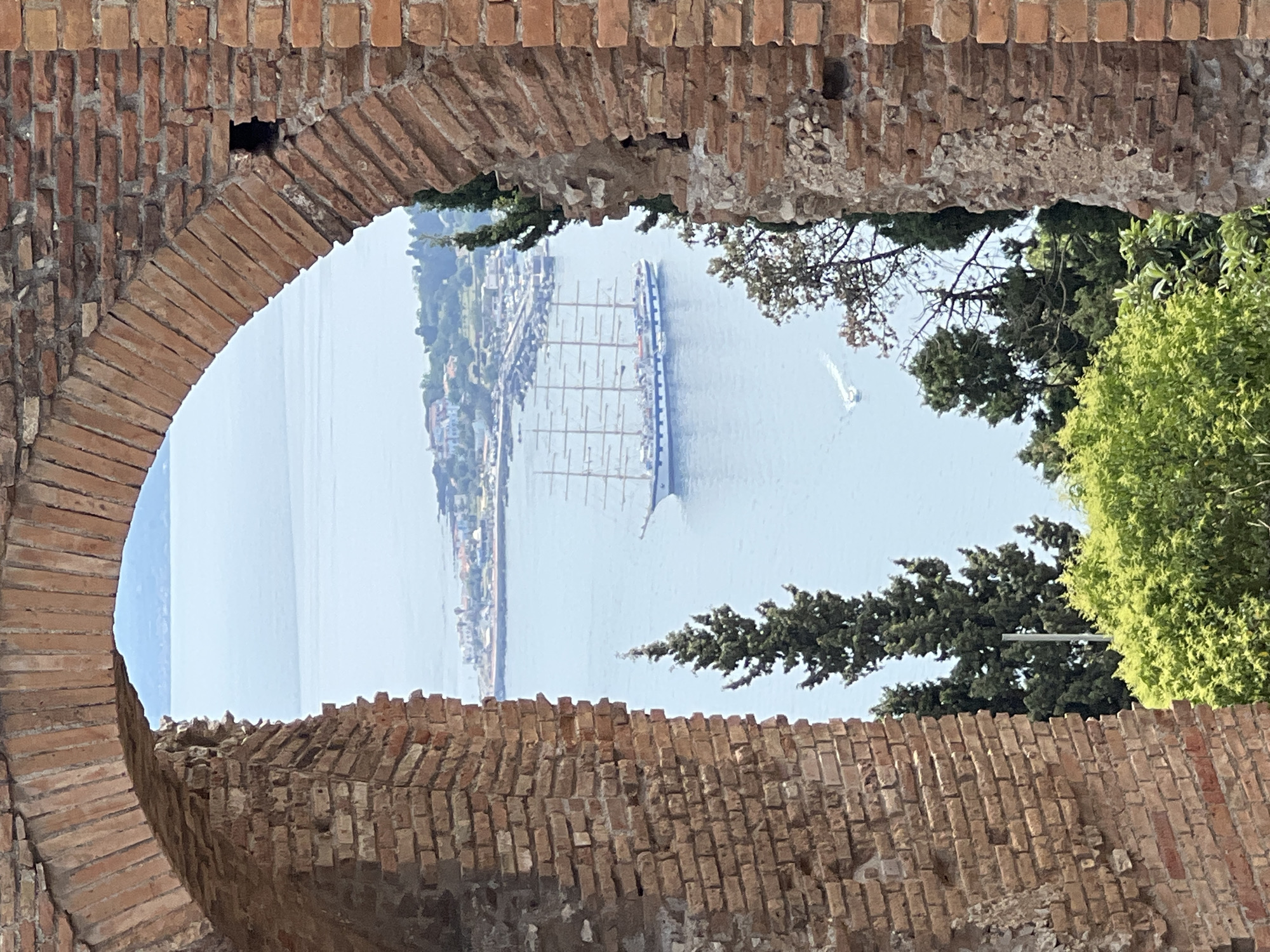 The image depicts a view through an ancient Roman archway in Taormina, Italy, overlooking the Ionian Sea. A large, multi-masted sailing ship is prominently featured on the water, appearing to be at anchor or moving slowly. In the background, a coastal town with buildings and a harbor can be seen. Lush green trees and vegetation frame the foreground and the lower portion of the arch. The arch itself is constructed of weathered red brick, showing signs of age and erosion. The sky is a light blue, suggesting a clear, sunny day. The scene captures a blend of historical architecture and natural beauty characteristic of Taormina.