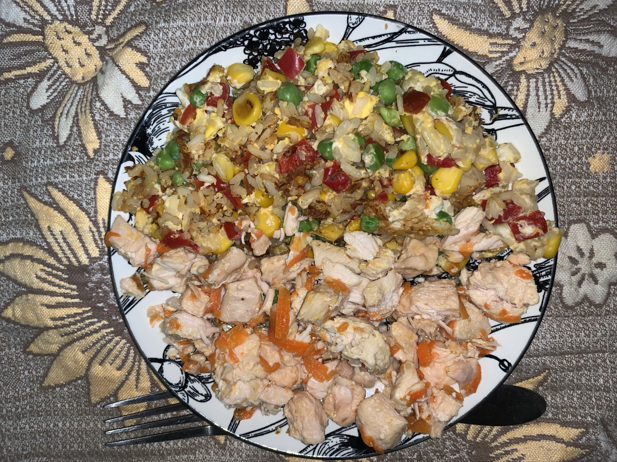 A top-down, close-up shot shows a plate of food. The plate is filled with a portion of what appears to be fried rice mixed with corn, peas, and diced red peppers, alongside a portion of what is seemingly chopped chicken, some pieces having an orange tint. A fork is positioned to the left of the plate, while a black object, possibly a knife handle, is visible to the right. The plate is set on a fabric with a floral pattern in shades of brown and gold. The city of Unknown, Unknown is not determined from this image.