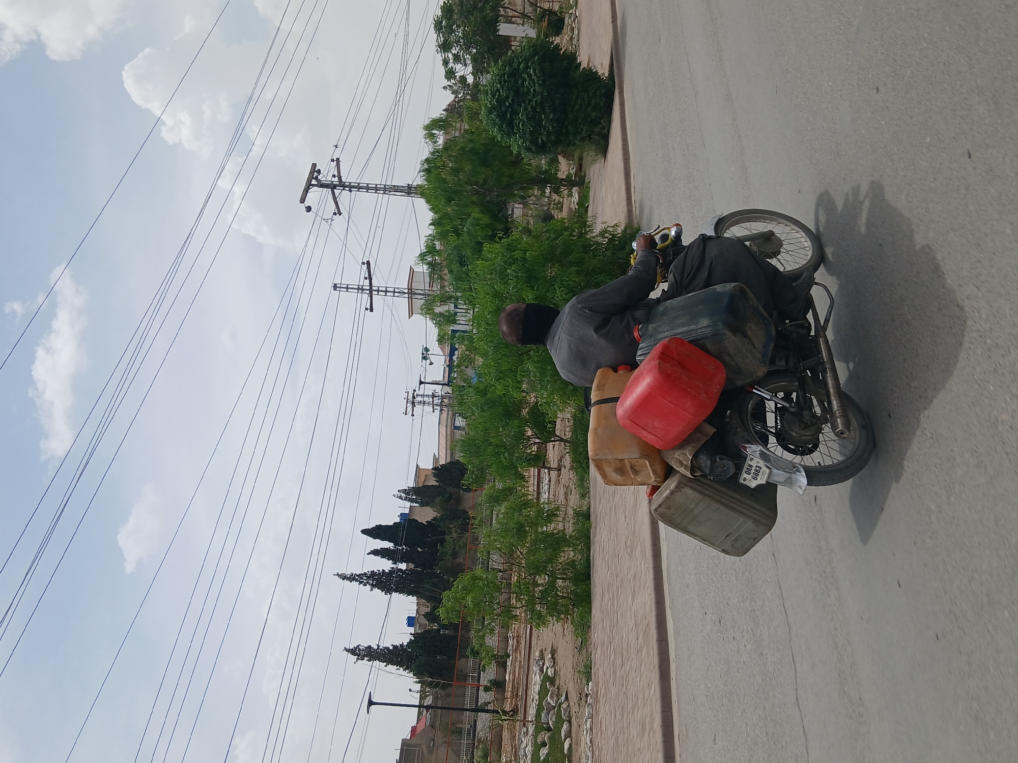 A man rides a motorcycle on a paved road in Quetta City Tehsil, Pakistan. The motorcycle is heavily laden with several plastic containers of varying sizes and colors. Overhead are utility poles with numerous electrical wires crisscrossing the sky. Lush green trees and bushes line the side of the road. The sky is a light blue with scattered white clouds. In the distance, buildings can be seen nestled amongst the trees.