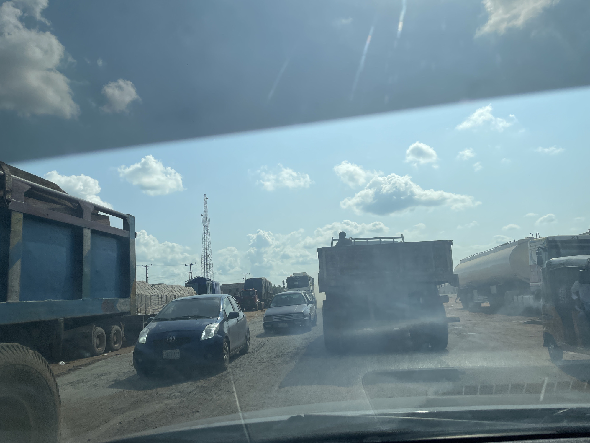 The media file depicts a traffic jam on a dusty, unpaved road in Adonkolo, Nigeria. The scene is dominated by a variety of large trucks, including dump trucks and a tanker truck, as well as smaller vehicles like a blue Toyota Yaris and a dark sedan. A three-wheeled vehicle is also visible. Several individuals are present, some appearing to be drivers or passengers, and one person is seen riding on the back of a truck. The road surface is dirt and appears dry and dusty, suggesting arid or dry weather conditions. The sky is partly cloudy, with a mix of dark and light clouds, indicating a dynamic weather situation. A tall transmission tower is visible in the background, a common landmark in many locations. The overall impression is one of congestion and slow movement, typical of a busy roadway in a developing area. The lighting suggests it is daytime, likely late morning or early afternoon. There are no discernible emotions conveyed by the subjects, as the focus is on the vehicles and the traffic situation. One license plate on the blue Toyota Yaris is partially visible, reading "LLH 74W".