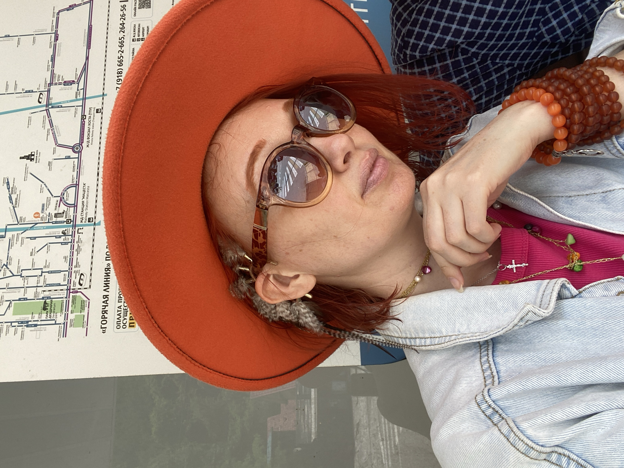 The image depicts a woman wearing an orange hat, sunglasses, and a denim jacket over a pink shirt, with multiple beaded bracelets and necklaces. She is posing in front of a map featuring the text "ГОРЯЧАЯ ЛИНИЯ" which translates to "HOT LINE" and "ЖД СТАНЦИЯ 'МАЦЕСТА'" which translates to "MAZESTA RAILWAY STATION". The map also contains the text "Ж/Д ВОКЗАЛ ХОСТА (ТПУ)" translating to "KHOSTA RAILWAY STATION (TPU)". The map also features the phone number "+7 (918) 665-2-665, 264-26-56". The area is likely in the city of Sochi, Russia, given the Russian language text.
