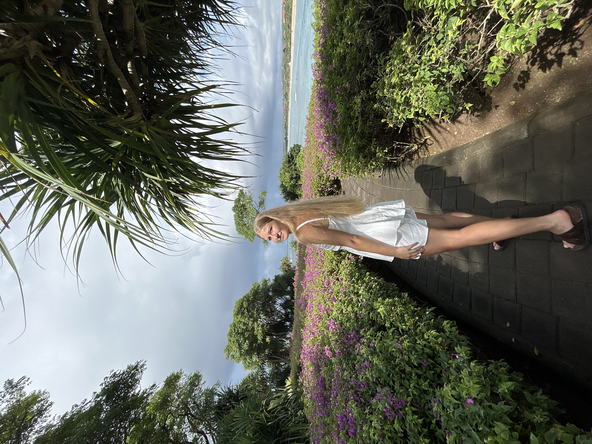 A young woman with long blonde hair, wearing a white sleeveless dress and brown sandals, stands smiling on a paved pathway in a tropical coastal setting. The pathway, constructed of dark grey blocks, leads through lush vegetation. To the woman's left, a vibrant border of green bushes interspersed with bright purple flowers (likely bougainvillea) extends into the background.
In the mid-ground, a serene light blue ocean stretches towards a distant shoreline, where faint outlines of buildings are visible. The sky above is partly cloudy, showing a mix of grey clouds and bright areas, indicative of daytime, possibly late morning or early afternoon. Large, spiky-leaved tropical trees frame the upper left foreground, adding to the lush environment. The woman looks directly at the camera with a happy and relaxed expression, appearing to pose for a photograph. The scene conveys a pleasant, sunny atmosphere in a scenic outdoor location, consistent with Kuta Selatan, Indonesia. There is no visible text in the image.