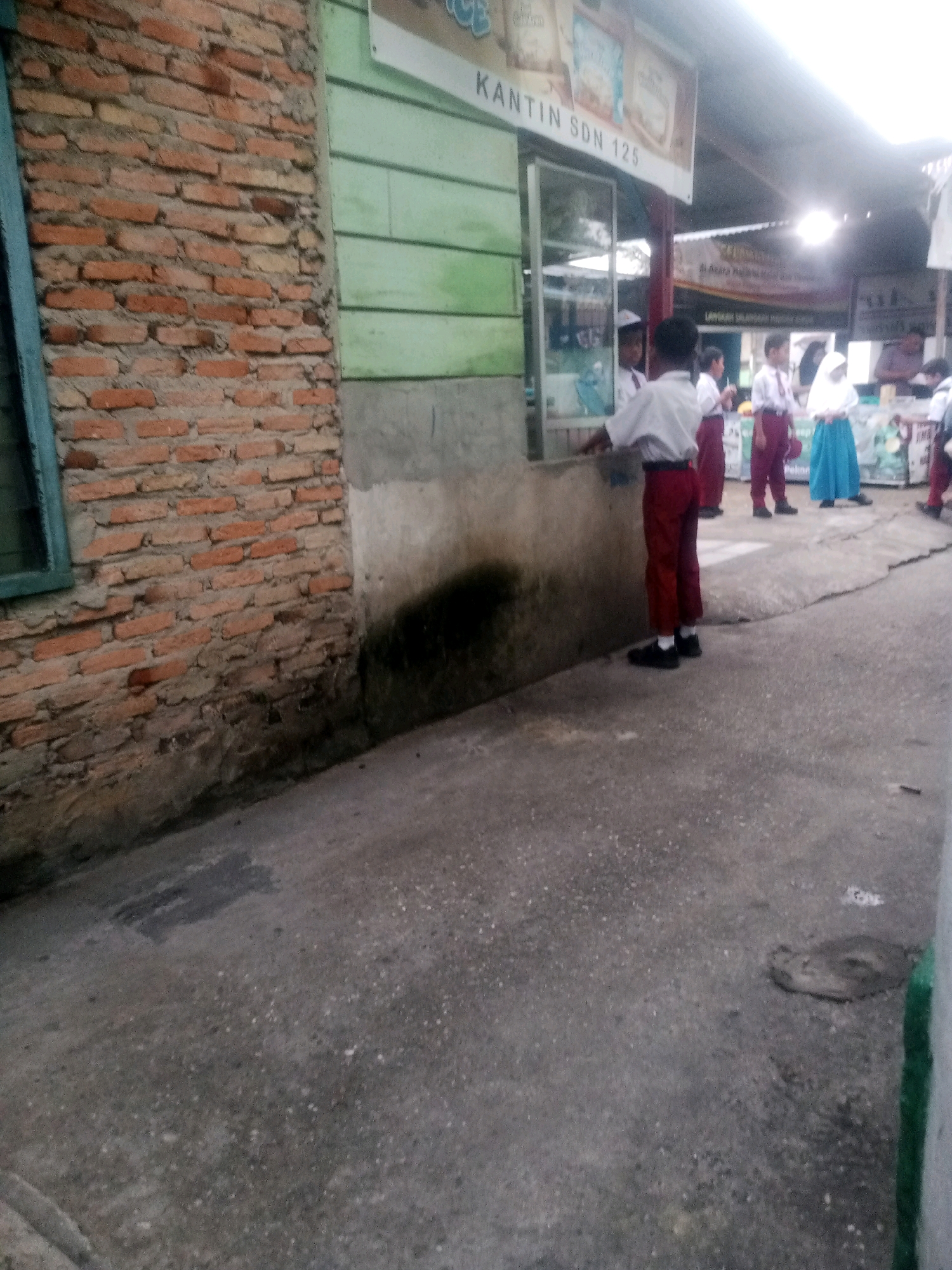 The image shows a scene in front of a canteen labeled "KANTIN SDN 125," likely located in Pekanbaru, Indonesia. Several children in school uniforms, consisting of white shirts and red trousers or skirts, are visible. One boy in the foreground, facing away from the camera, is wearing a white shirt, red trousers, and black shoes. He appears to be interacting with someone inside the canteen through a window. Other children are standing or walking in the background. The building has a brick wall on the left and a green wooden wall with a window. The ground is paved with concrete. The overall environment suggests a schoolyard or an area adjacent to a school.