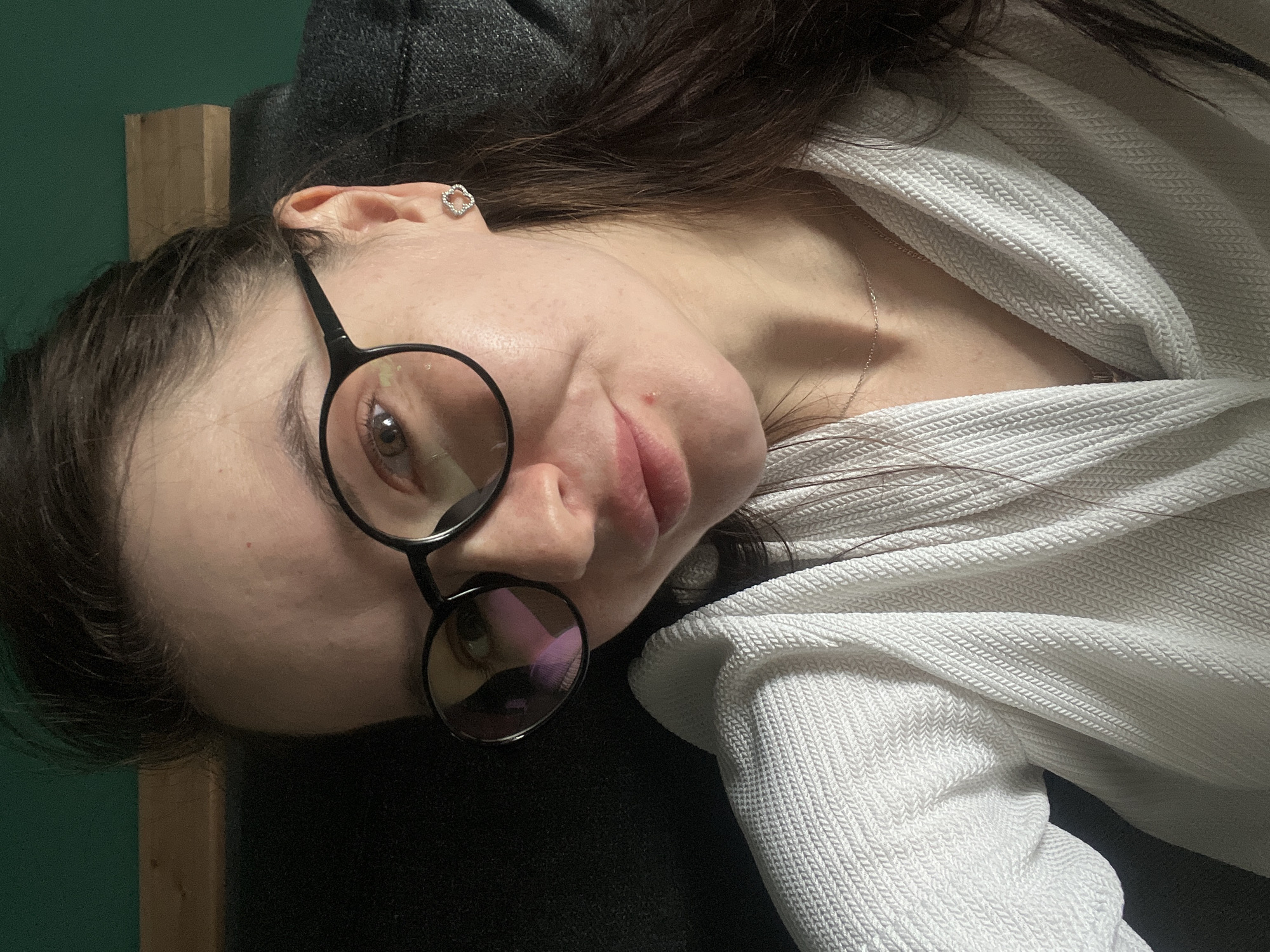 A woman with dark hair and glasses is angled slightly towards the viewer. She is wearing a white knit shirt and a delicate silver necklace. She has an earring in her left ear, appearing to be a small, square-shaped design. A wooden beam is visible behind her head, and a dark green wall is partially visible in the background. A black object appears near her hair, possibly a piece of furniture. The location cannot be identified as Kazan, Russia from the information in the image.