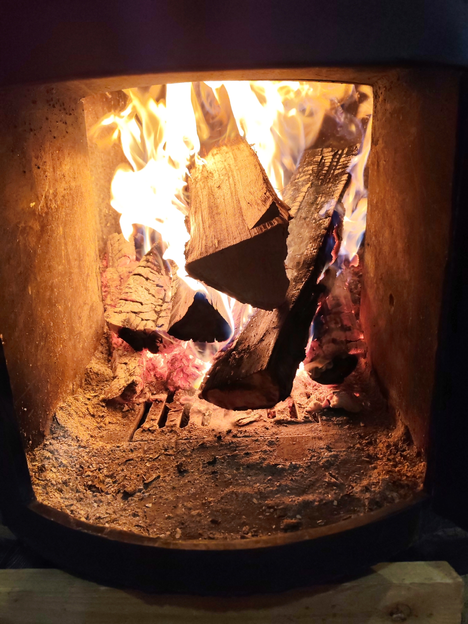 The image shows a close-up view into a lit fireplace. Inside, logs of wood are burning, with visible flames and embers. The interior of the fireplace is lined with a rough, tan-colored material. The floor of the fireplace is covered with ash and wood debris. There is no evidence in the image to suggest a location in the city of Chaikovsky, Russia.