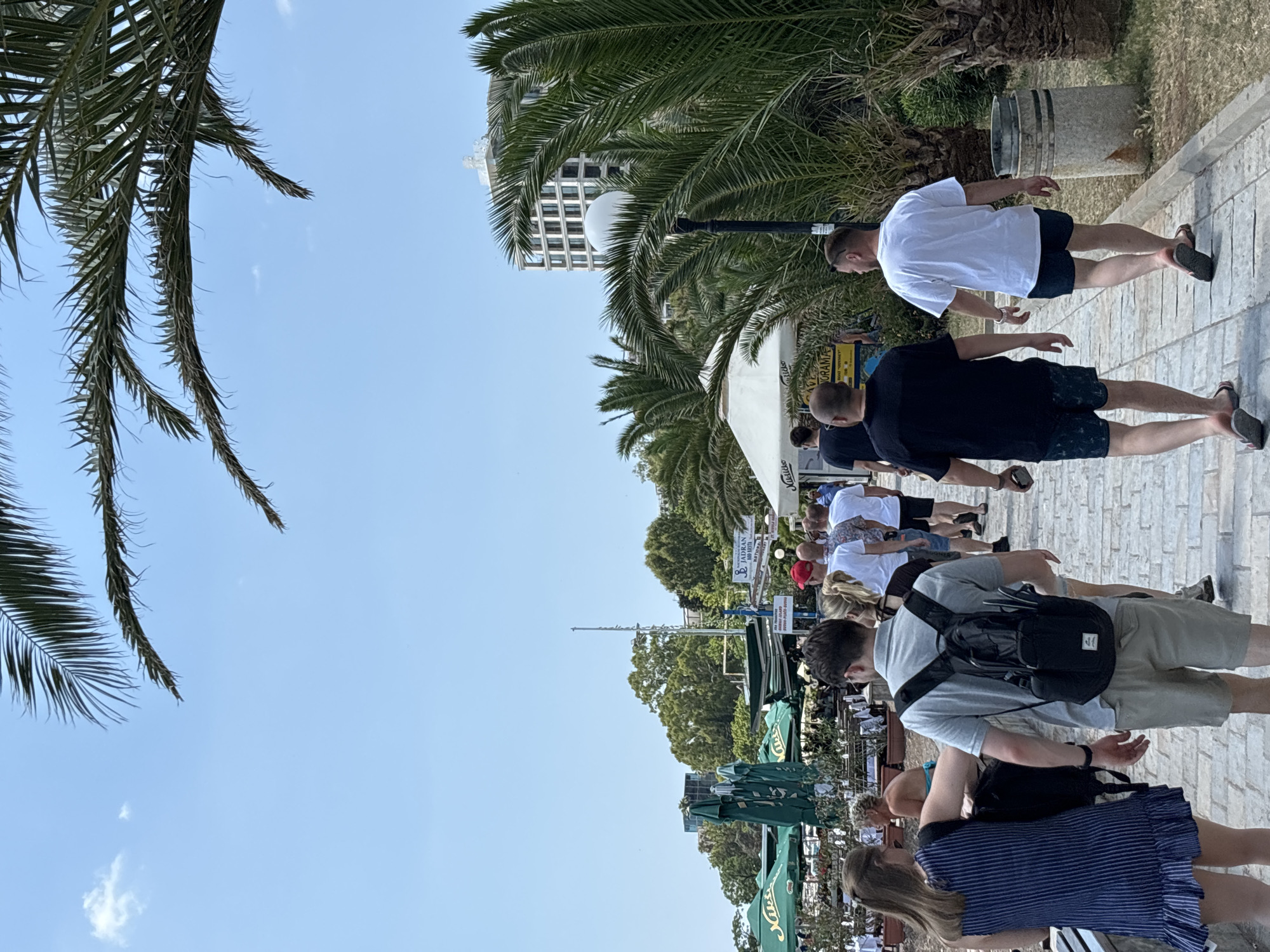 A group of people walk along a paved pathway in Budva, Montenegro, on a sunny day. Palm trees line the right side of the path, and a building with a grid-like facade is visible in the background. Several green umbrellas, likely for outdoor seating at a restaurant or cafe, are visible on the left side of the image, along with tables and chairs. The people are dressed in casual summer clothing, including shorts, t-shirts, and a dress. One person in the foreground wears a blue and white striped dress, and another wears a grey polo shirt and khaki shorts, carrying a black backpack. The pathway is made of light-colored paving stones. The sky is clear and blue.