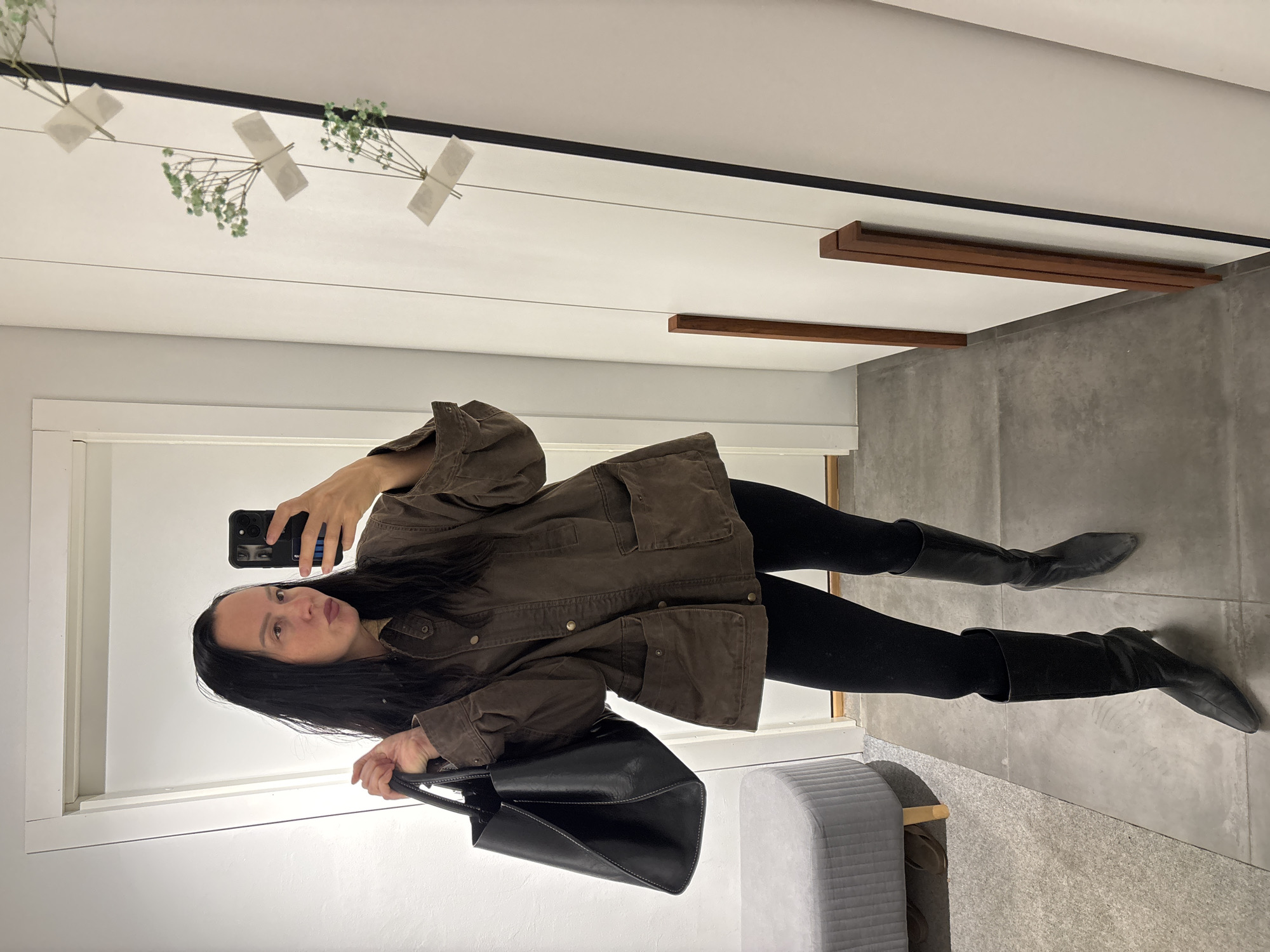 A person with long dark hair is taking a selfie in a room. They are wearing a brown jacket, black pants, and black boots. They are holding a black purse. The room has white walls, a gray floor, and some decorative elements on the ceiling. The location is not specified in the image and cannot be determined.
