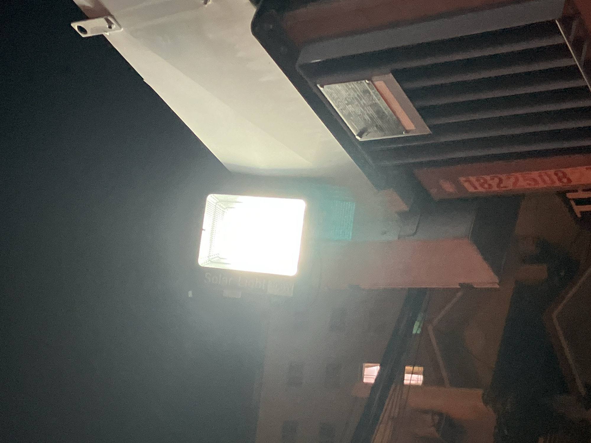 A bright solar-powered LED floodlight is mounted on a pole in front of a building at night in Nha Trang, Vietnam. The floodlight illuminates a vertical sign on the pole which reads "Solar Light". To the right of the floodlight is a portion of a white building. Below the floodlight and to the left, a multi-story building with illuminated windows is visible. In the foreground on the right, a dark metal fence with vertical slats runs along the side of a shipping container. A metal plate is affixed to the fence. The shipping container has orange markings on its side, including the number "1827508". The sky is dark.