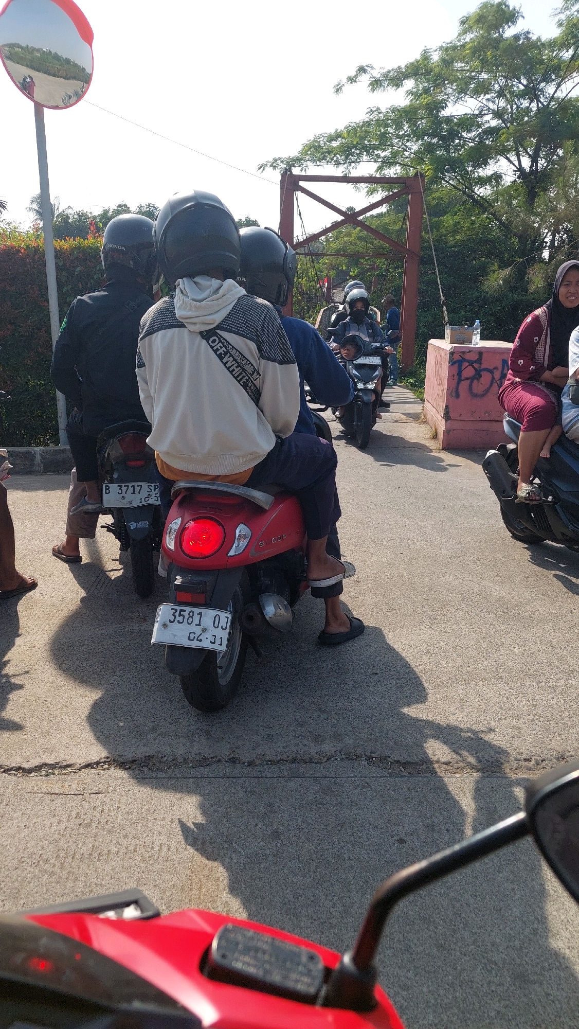A group of people on motorcycles are stopped on a paved road in Cibinong, Indonesia, likely waiting to cross a bridge. The focus is on a red scooter in the foreground, with its brake light illuminated. The scooter has a license plate that reads "3581 OJ". A person wearing a white hooded sweatshirt with "OFF-WHITE" printed on the back is seated on the scooter. Behind them, two other individuals wearing helmets are on another motorcycle. In the background, more motorcycles and people are visible, along with a red metal bridge structure and lush green trees. A convex traffic mirror is mounted on a pole to the left of the frame. The scene suggests a warm, sunny day.
