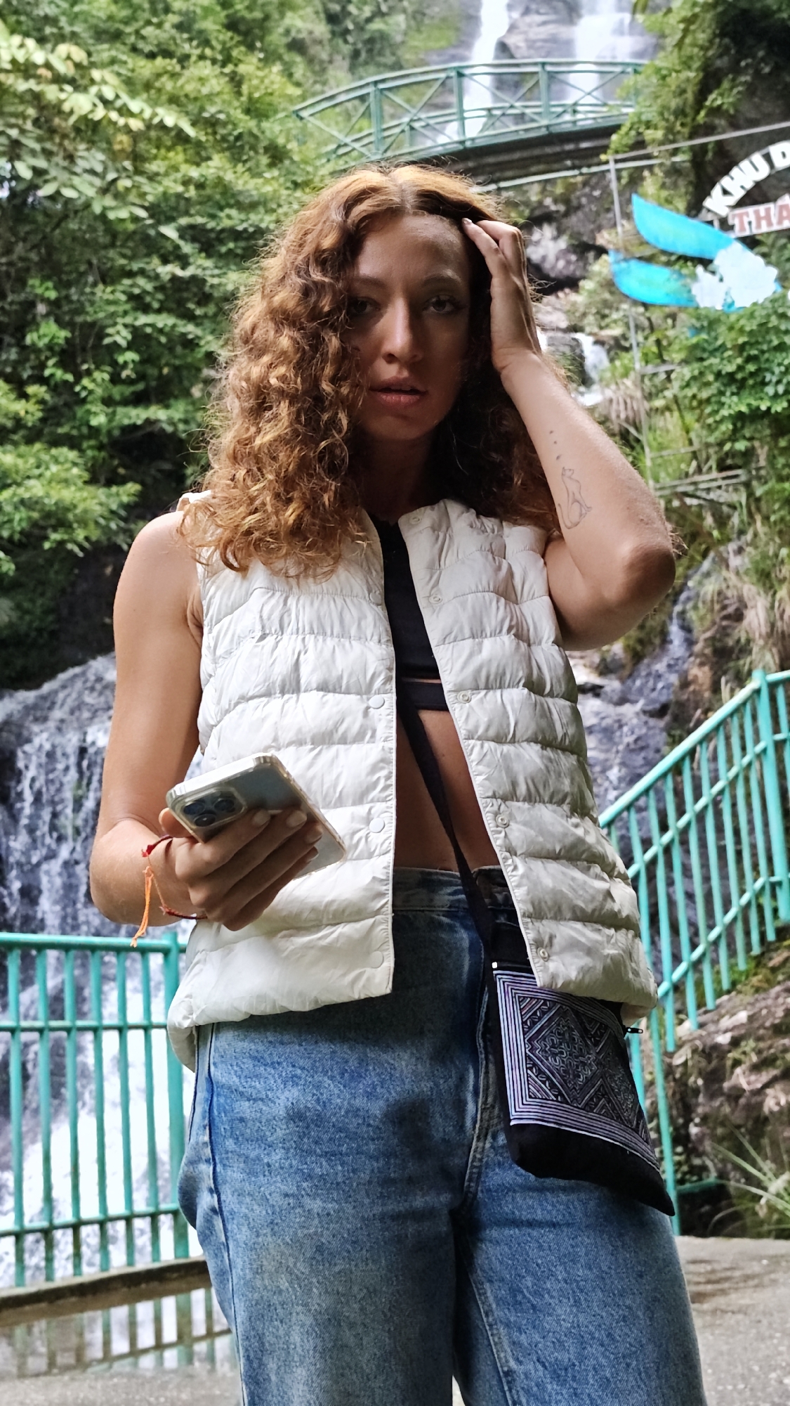 A person stands in front of a turquoise railing with a waterfall in the background. The person has light brown curly hair, and is wearing a white vest, blue jeans, and a black bandeau top. They have a small black bag hanging from their left side. They are holding a smartphone in their left hand and their right hand is touching their hair. The background shows a rocky landscape with lush green vegetation, and a bridge across the waterfall. The location is Sa Pa Ward, Vietnam.