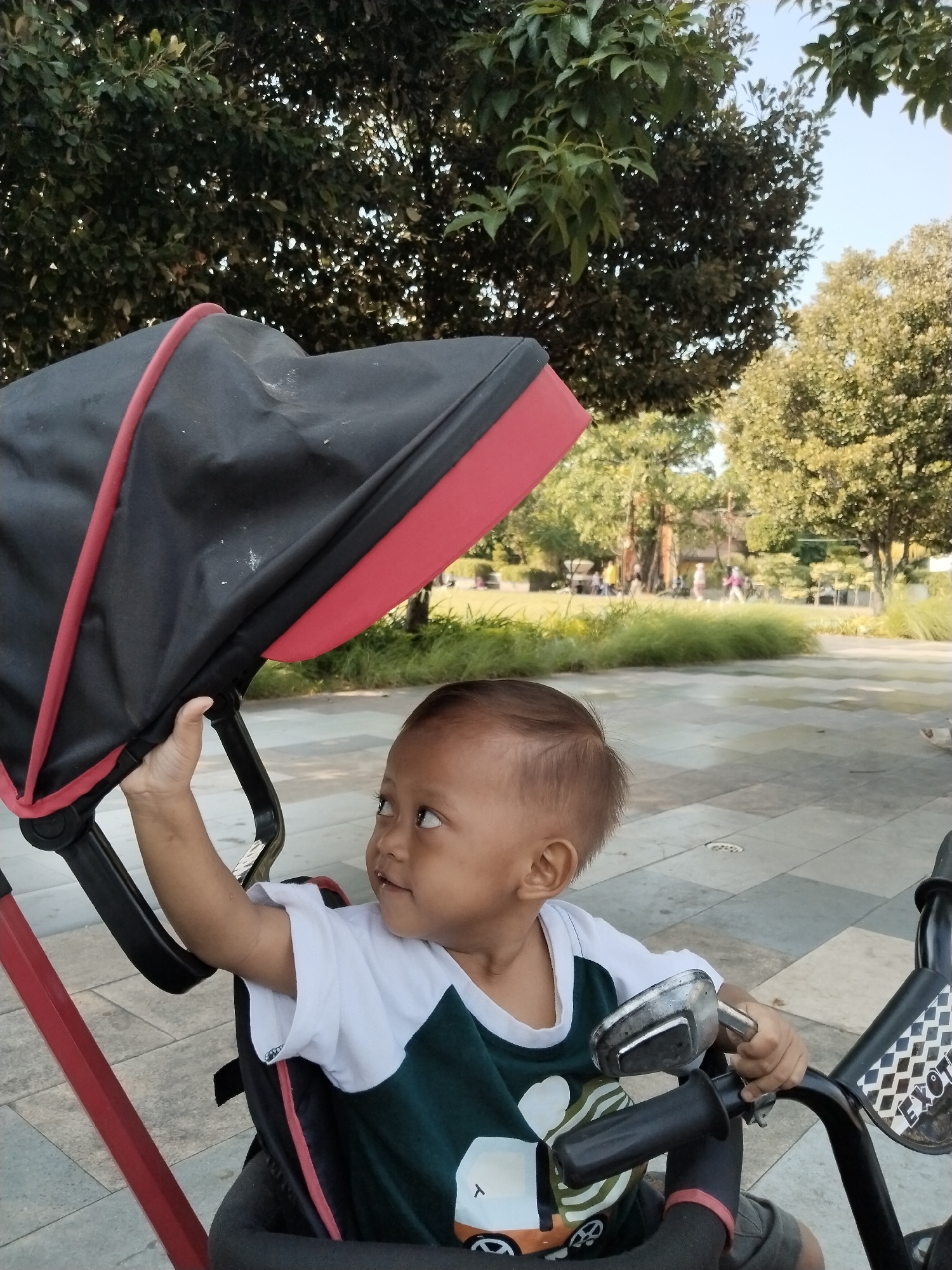 A young child sits in a stroller in an outdoor paved area, with trees and greenery in the background. The child is wearing a white t-shirt with a dark green graphic design. The child is reaching up with their right hand, holding onto the stroller's canopy. The stroller has a black canopy with red trim and a red frame. The ground is paved with light gray and beige rectangular tiles. In the background, there are people walking and trees. The sky is visible and appears clear and blue. The overall scene suggests a public park or recreational area in Kecamatan Pati, Indonesia.