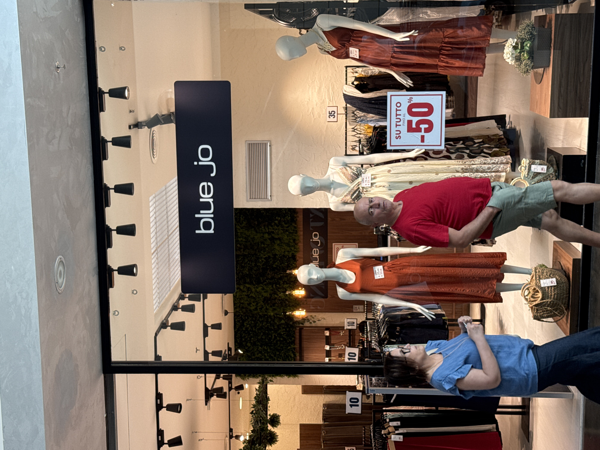 In Carini, Italy, a storefront window displays mannequins adorned in dresses, with sale signage indicating "SU TUTTO FINO AL -50%". A man in a red shirt and green shorts walks past the window, while a woman in a blue shirt and dark pants stands to the left, looking towards the store. Racks of clothing are visible inside the store, along with decorative greenery and lighting fixtures. The store's name, "blue jo", is displayed on a prominent sign above the window. A wicker basket with greenery sits on a wooden pedestal in the foreground.