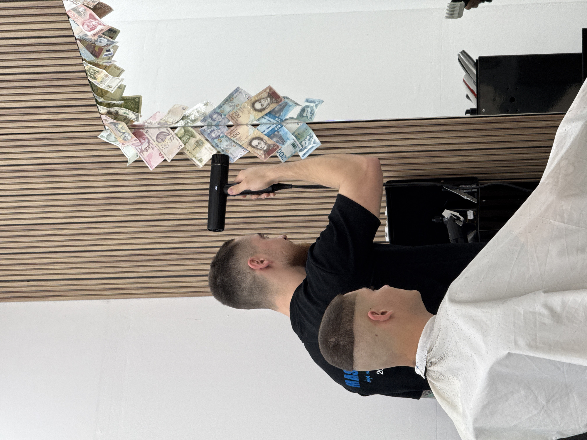 A barber in a black t-shirt is providing a haircut to a client in a white barber's cape. The client is turned to the right. The barber is using an electric hair trimmer. The room has a mirror, framed by a light-colored wooden plank backdrop, which is decorated with various international banknotes. The walls are painted white. The location is likely Sofia, Bulgaria, given the presence of Bulgarian banknotes in the mirror decoration.