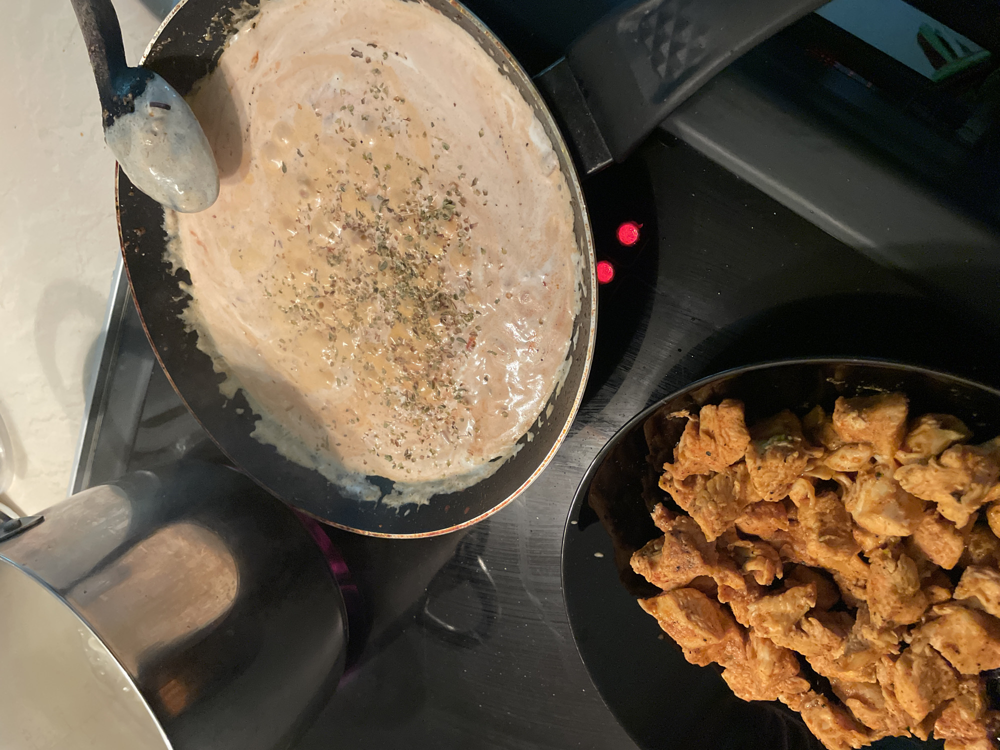 This is a close-up, high-angle, eye-level shot of a stovetop cooking scene. The image primarily features a frying pan with a creamy, pinkish sauce being stirred with a light-colored spatula. Dried herbs are sprinkled on top of the sauce, and the sauce appears to be simmering. To the left of the frying pan, a black bowl is filled with bite-sized pieces of seasoned, cooked chicken. In the background, a large, dark pot with a metallic sheen sits on a burner, emitting a faint purple glow from the heat. The stovetop is black and appears to be an induction or glass-ceramic cooktop, as indicated by the red indicator lights. The scene is indoors, likely in a kitchen. The location in Zlatograd, Bulgaria cannot be determined from the image.