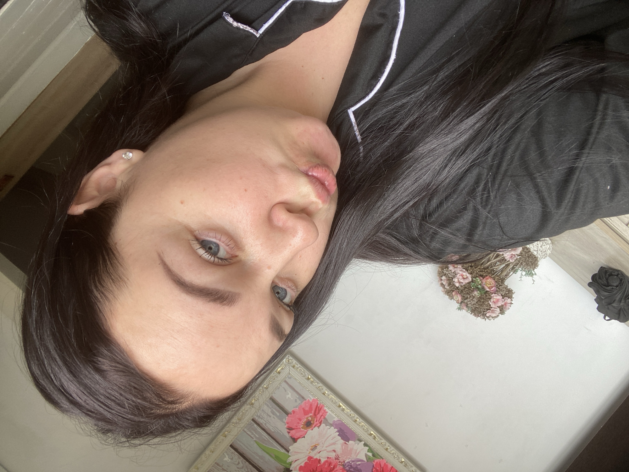 A woman with blue eyes and dark hair is facing the camera. She is wearing a black top with white trim. Her lips are pursed. She has an earring in her left ear. A picture with pink and white flowers is in the lower left corner. A flower bouquet and a black object are in the upper right corner on a white surface. The location and city cannot be determined from the visual information provided.