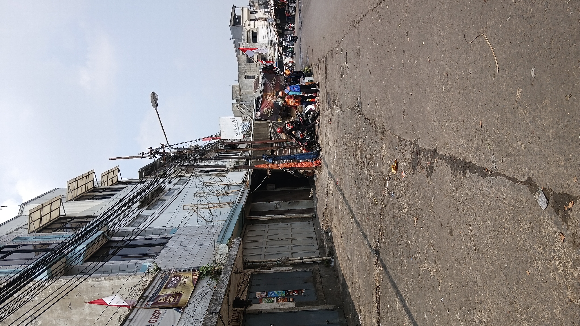 The image depicts a street scene in Bandung City, Indonesia, characterized by a narrow road with worn asphalt and scattered debris. On the left, a multi-story building with a light-colored facade features multiple windows covered by angled awnings. Wires are strung haphazardly across the building's exterior. A small Indonesian flag is visible on a pole attached to the building. Several closed storefronts with grey metal roll-up doors line the sidewalk. Further down the street, on the right side, several individuals are gathered near a motorcycle and a makeshift stall covered by a banner. One person wearing a red helmet and black clothing is on the motorcycle, while others in casual attire stand nearby. In the background, more buildings are visible, along with other motorcycles and people. The sky is overcast with light clouds. The overall impression is of a busy, perhaps commercial, area with a somewhat unkempt appearance.
