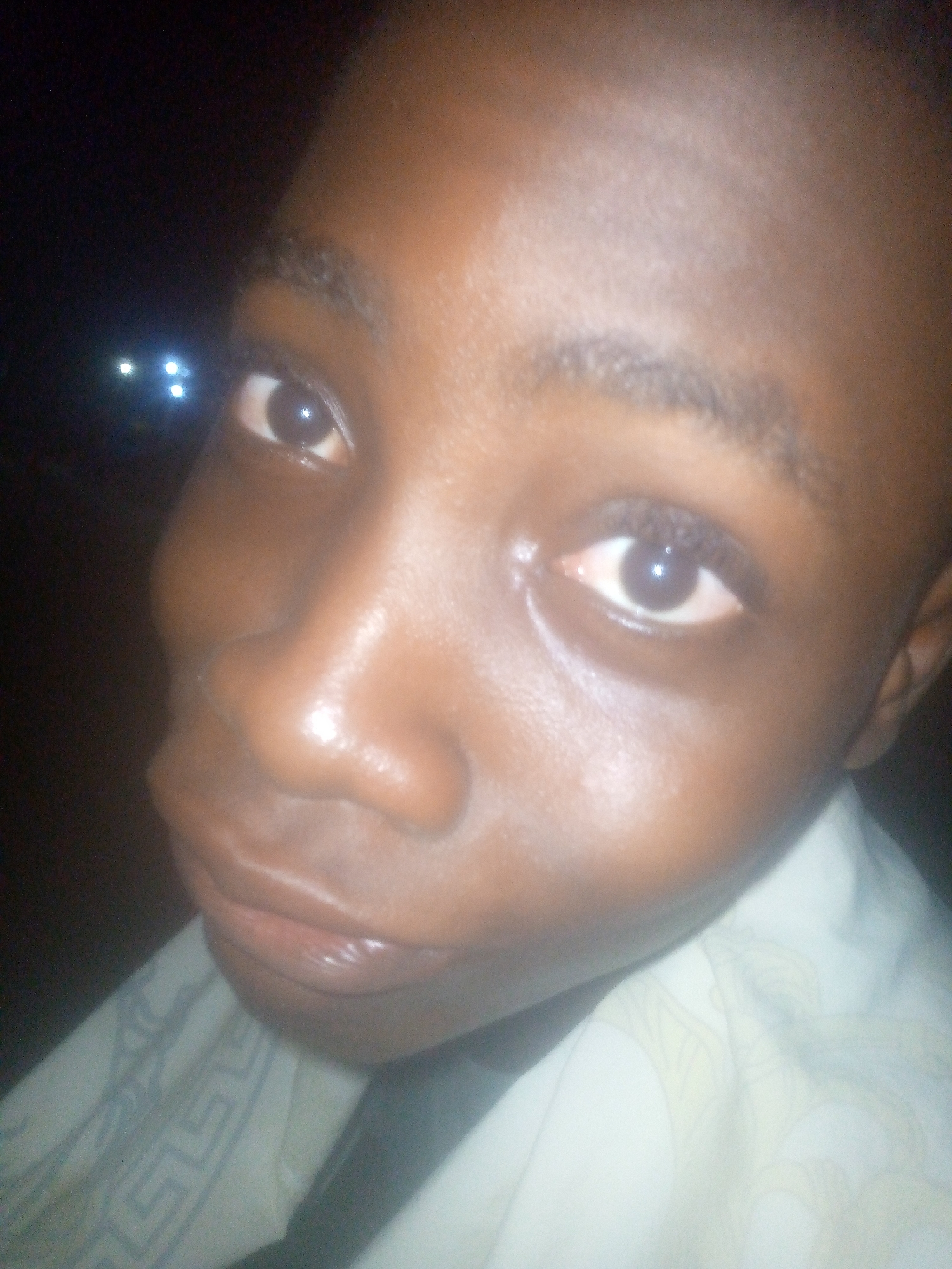 A close-up image shows a person with dark skin. The person is wearing a light-colored patterned shirt. The location appears to be outdoors at night, with blurred lights visible in the background. The city and state are unknown.