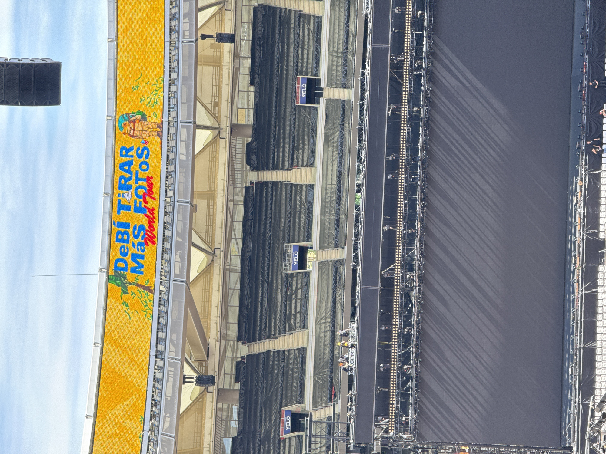 An aerial view captures the interior of a large outdoor stadium or arena, oriented vertically as if looking down from a high vantage point. The sky, a clear blue, occupies the top portion of the frame.
Along the upper edge of the stadium structure, a prominent, elongated LED screen or banner displays vibrant yellow, textured background. On this banner, blue text reads: "¡DeBi TIRAR Más FOTOS!" followed by "World Tour" in a smaller, red font below. An illustrated character, resembling a stylized, possibly frog-like figure wearing a hat and carrying a camera or backpack, is depicted next to the text.
Below the banner, several seating sections are visible, largely covered with dark, protective tarps, indicating they are either unoccupied or being prepared for an event. Interspersed among these sections are small rectangular screens displaying the brand name "VELO" in white letters against a blue and red background. Structural elements of the stadium, including beams and scaffolding, are evident throughout this mid-section.
The lower half of the image features a vast, dark gray or black rectangular area, which appears to be the main stage or performance floor. Along the left and right edges of this stage, extensive arrays of lighting equipment and speakers are set up, casting diagonal shadows across the stage surface, suggesting a bright, sunny day. Several small figures of people, likely crew members, are visible on and around the stage, working on the setup.
The scene depicts the extensive preparation for a major event, such as a concert or festival, with clear skies overhead and the meticulous arrangement of stage equipment and venue branding.