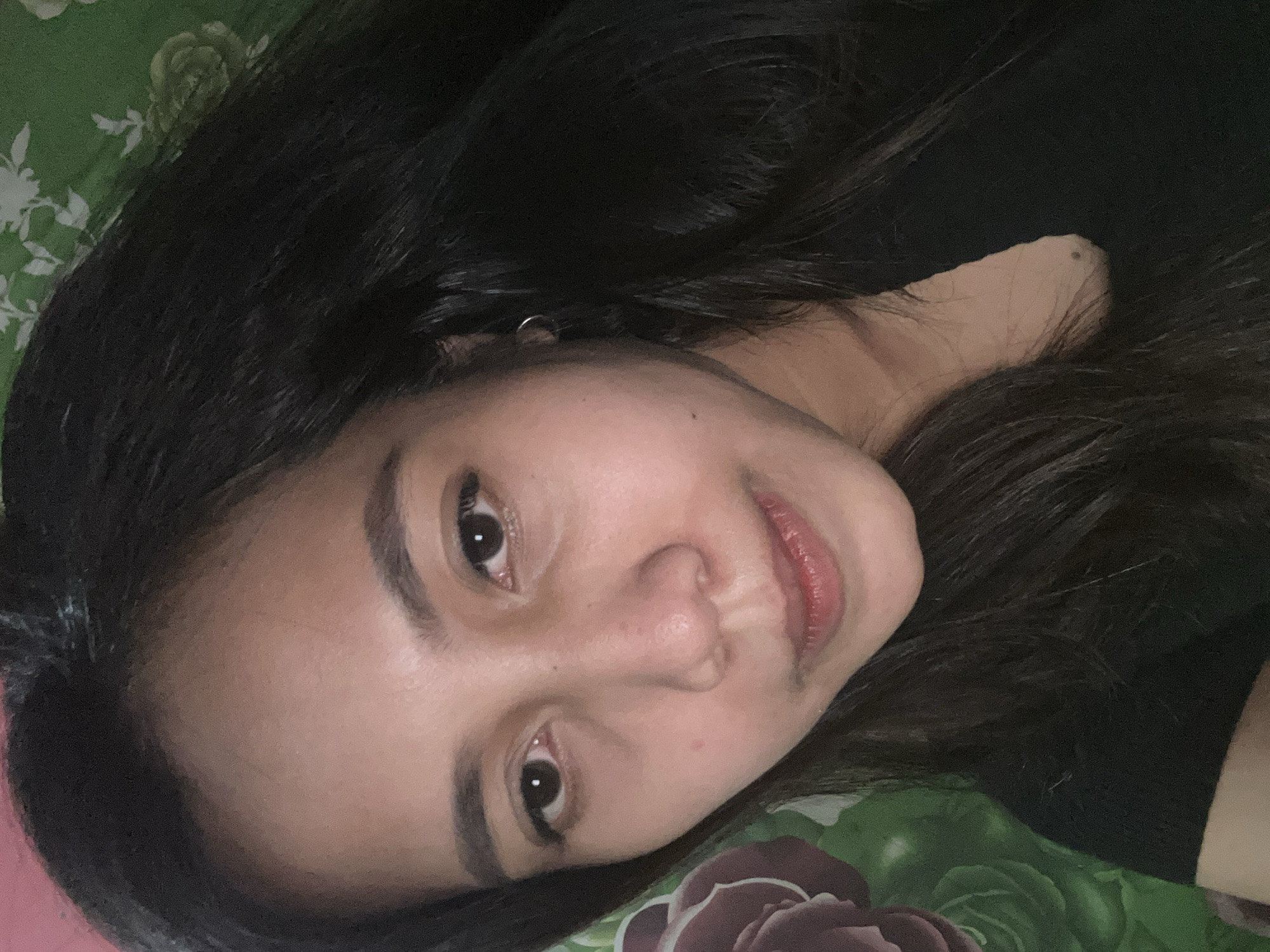This is a close-up, self-taken photograph of a young adult woman's face. She has long, dark hair that frames her face and is wearing a black top. Her skin tone is medium, and her eyes are dark brown. She has a slight smile. The background appears to be a patterned fabric, possibly a bedsheet or curtain, with green and floral motifs, suggesting an indoor setting. No specific landmarks or elements definitively place the image in Depok, Indonesia, but the general context is a casual personal photo.