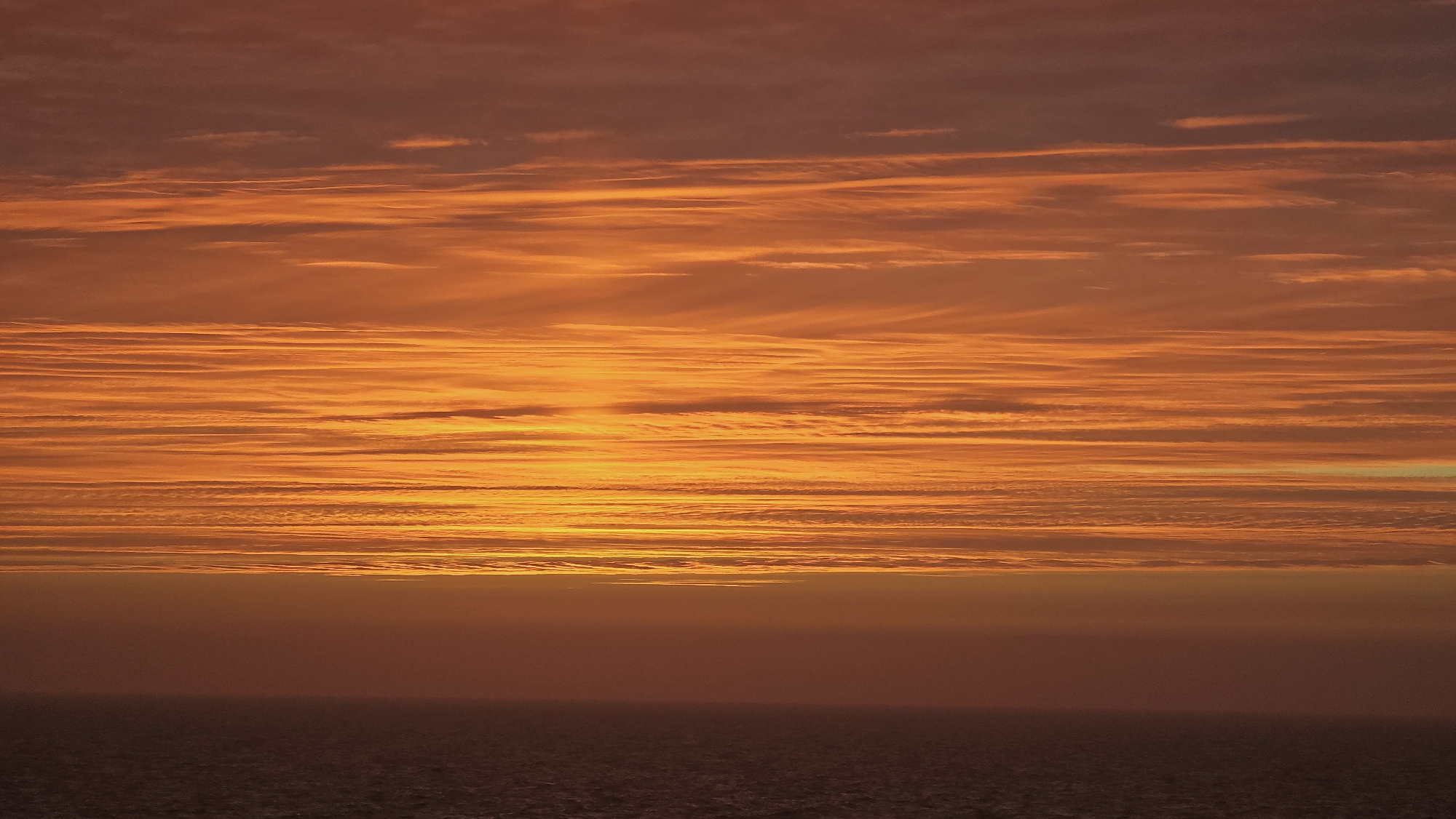 The image depicts a vibrant sunset over the ocean in an unknown location. The sky is filled with layers of orange and red altocumulus clouds. The horizon line is visible with a dark, murky ocean beneath the brightly colored sky. There are no people visible in the image.