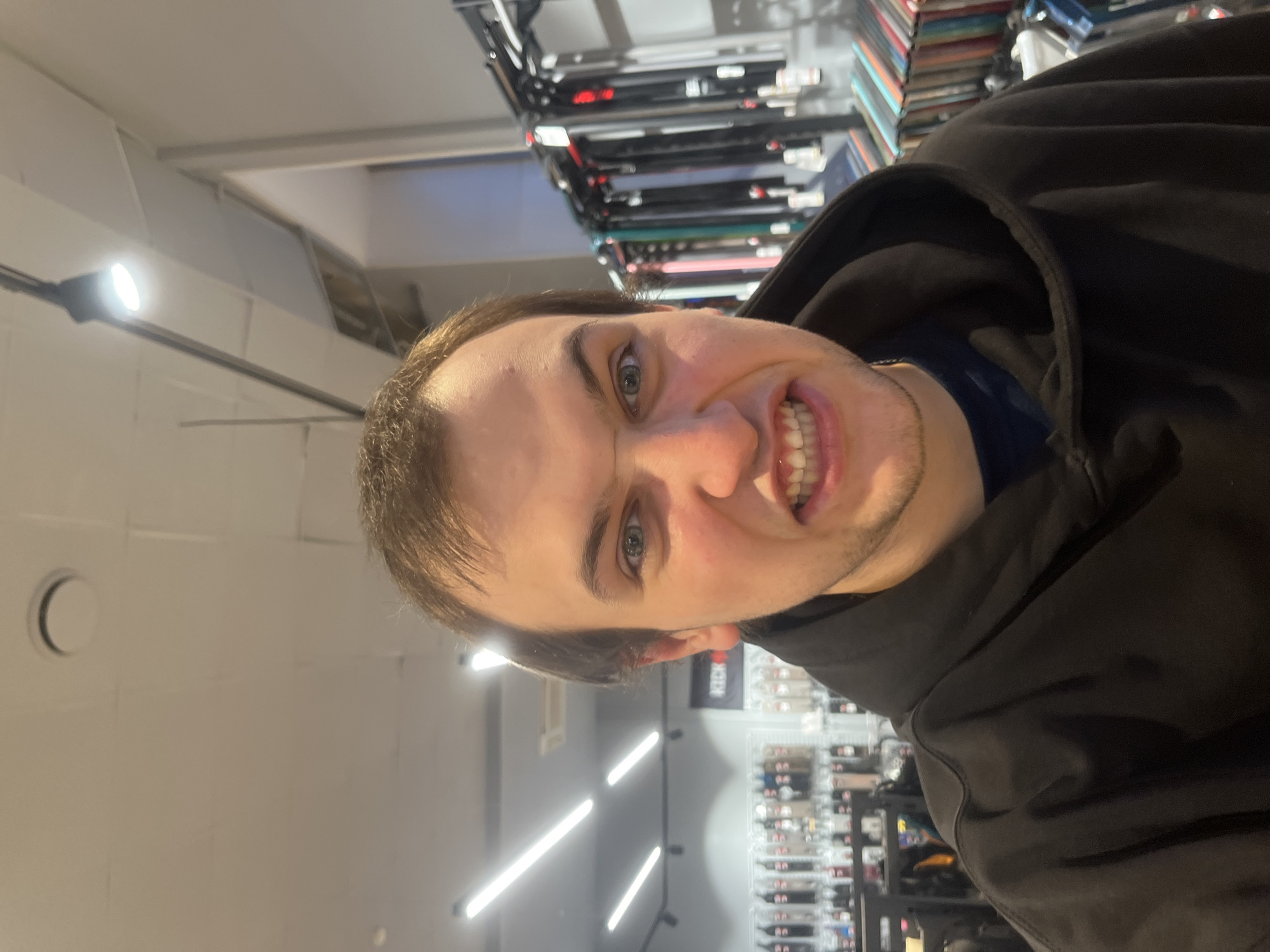 A man with short brown hair and blue eyes is grimacing at the camera. He is wearing a black hooded sweatshirt. The background appears to be a store, possibly in Krasnoyarsk, Russia. There are rows of products visible on shelves. The lighting is bright, with fluorescent lights illuminating the scene. The photo is taken at an angle.