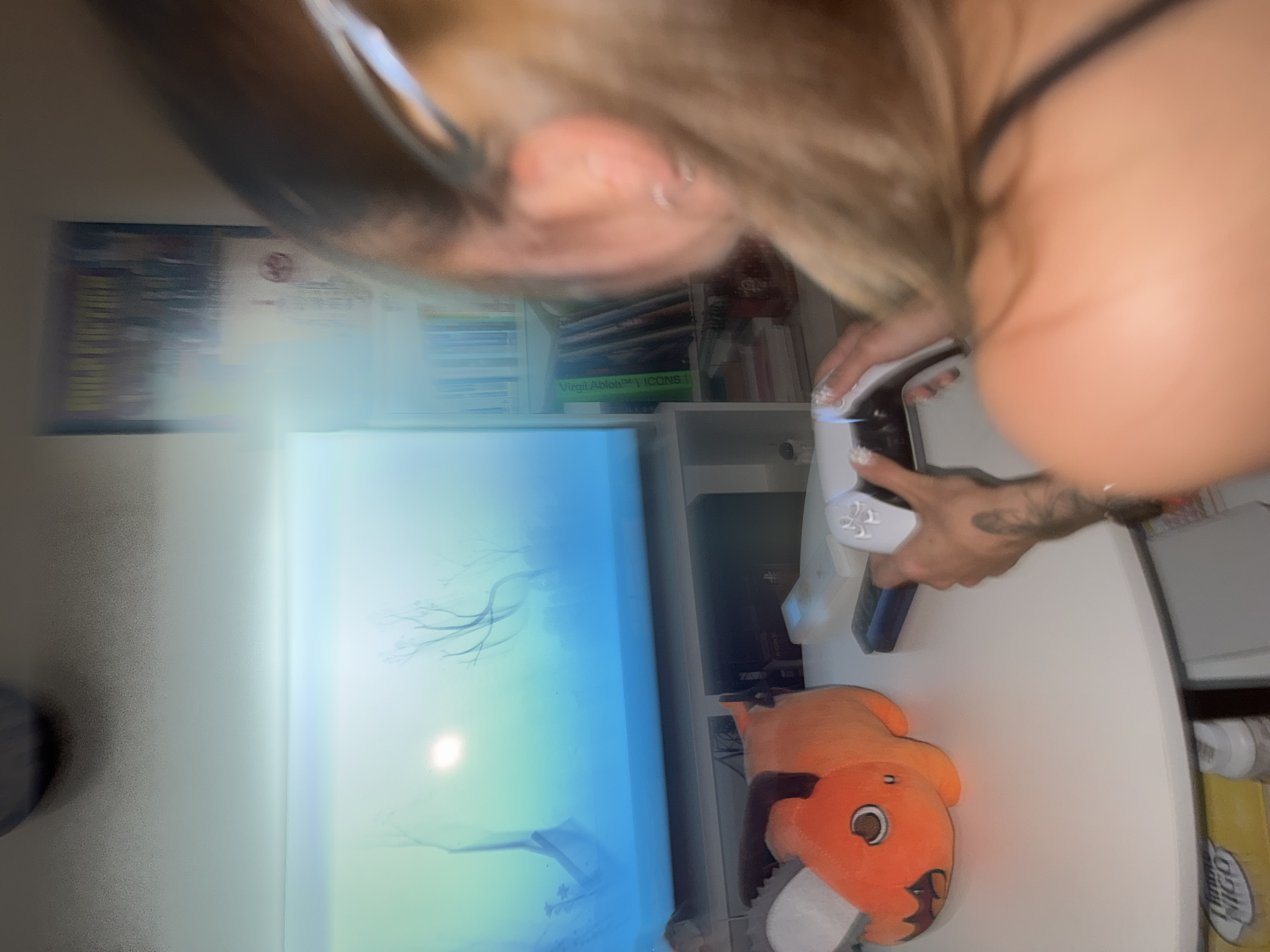 The image shows a person playing a video game in what appears to be a home environment. A large television screen displays a serene, possibly nature-themed, video game scene with a bright light source in the upper left quadrant and silhouetted trees. In front of the television, on a white table, is an orange plush toy that resembles a character from an anime or video game, and two gaming controllers. One controller, a white PlayStation 5 controller, is in the hands of the person. The person, whose back is to the camera, has long blonde hair and is wearing a black tank top. A bookshelf filled with books and what appear to be DVDs or video game cases is visible in the background, to the right of the television. The overall lighting suggests an indoor setting, likely during the day given the bright screen and ambient light. No specific location in Moscow, Russia can be determined from the visual information provided in the image.