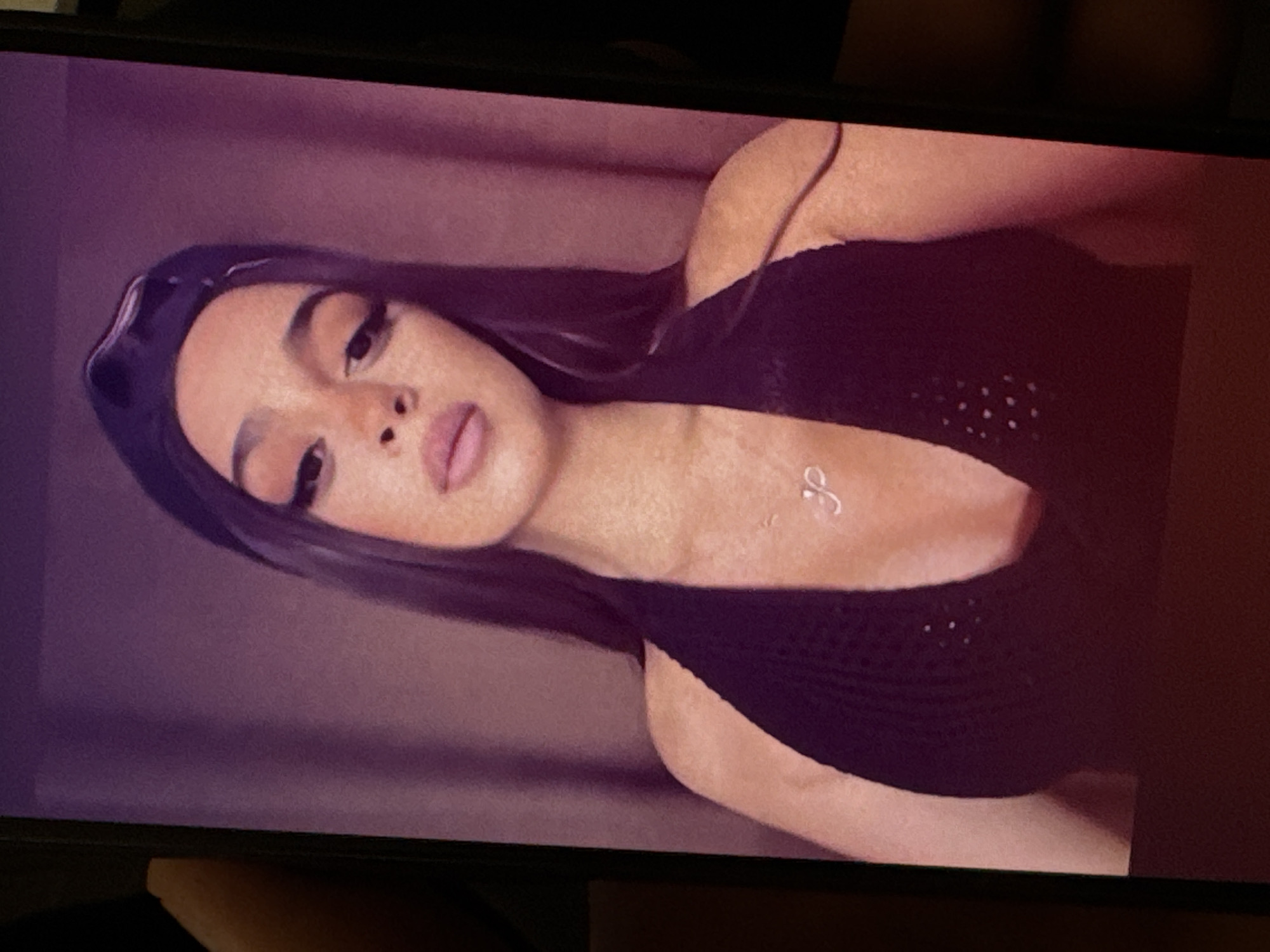 A woman with dark hair and heavy makeup is the main subject of the image. She is wearing a dark, possibly black, top with a thin strap, and a headband. There is a small, delicate necklace visible on her chest. The background is a blurred mix of purple and beige, possibly a curtain or wall. There is no discernible information to determine the image was captured in the city of Yerevan, Armenia.