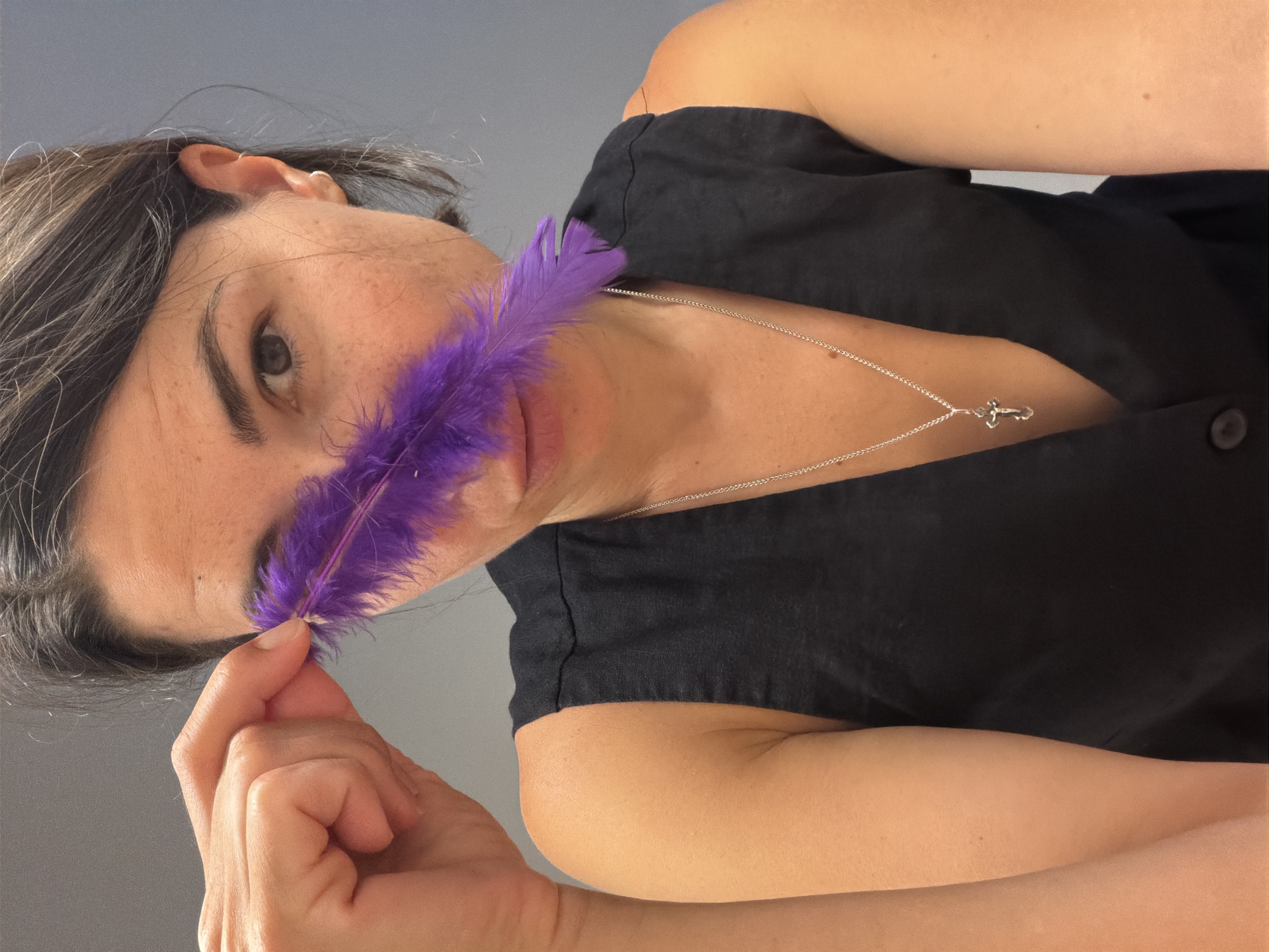 A woman is featured in a close-up, rotated portrait, likely taken indoors in Budva, Montenegro. She is looking directly at the camera with a neutral expression. Her head and upper torso are visible, against a plain, light grey background.
The woman has dark hair with some gray streaks, fair skin with visible freckles across her nose and cheeks, and dark eyebrows and eyes. She wears a black sleeveless top or dress, and a silver chain necklace with a small silver cross pendant rests on her chest. Her right hand, with neat fingernails, is holding a vibrant purple feather, positioning it near her nose and cheek. The lighting suggests an indoor setting, and no specific time of day or weather is indicated. There is no visible text in the image.