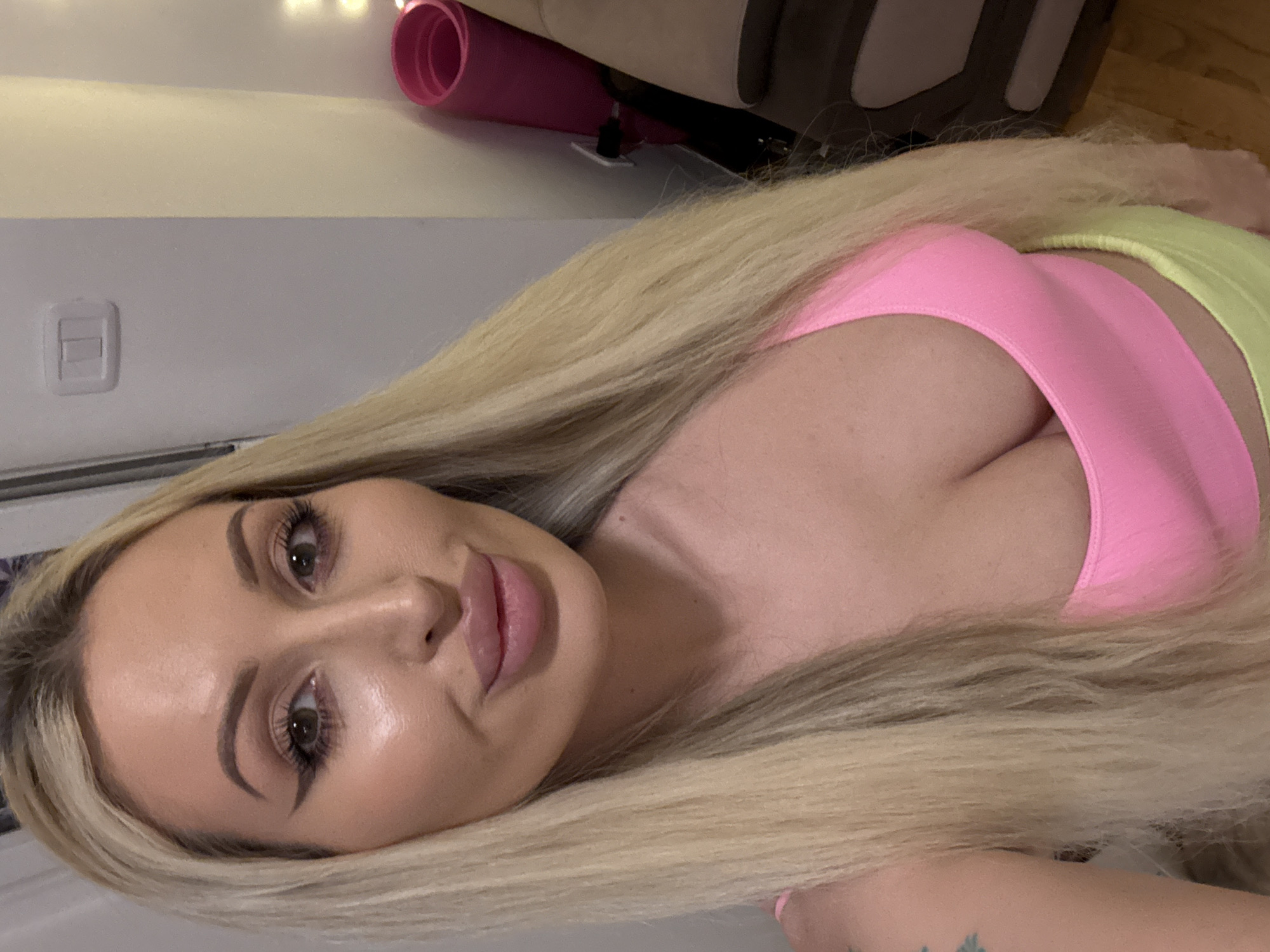 A woman with long blonde hair and prominent makeup, including winged eyeliner and full lips, poses for a selfie. She is wearing a pink crop top and a yellow skirt. The background shows a wall with a light switch and a rolled-up pink mat, possibly a yoga mat, leaning against it. A piece of furniture, possibly a couch, is visible on the right. The scene appears to be indoors.