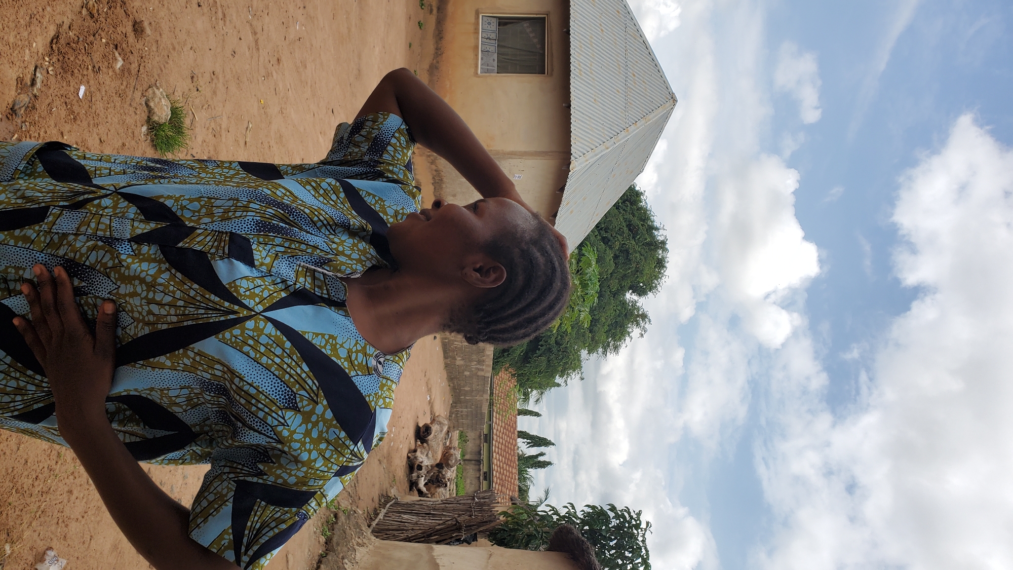 A medium shot captures a woman in profile, looking to her left, with her right hand touching her head and her left hand resting on her stomach. She is wearing a brightly colored patterned shirt with a predominantly blue, green, and black design. Her hair is styled in cornrows. The background depicts a rural or semi-urban setting in Jalingo, Nigeria. To the left, a light-colored building with a corrugated metal roof and a window with curtains is visible. Behind the woman, a dirt path leads into a courtyard. Further in the background, more buildings with tiled roofs and wooden fences can be seen, along with lush green trees. The sky above is partly cloudy.