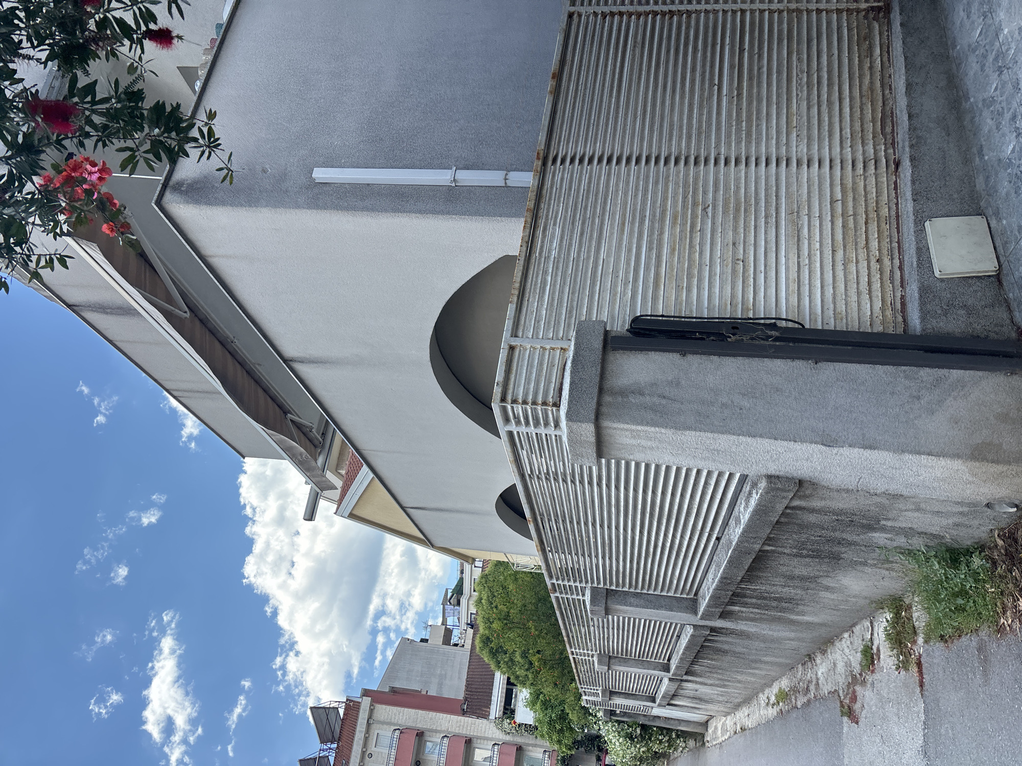 The image shows a street scene in Budva, Montenegro. The architecture features a mix of building styles, including a light-colored building with a curved archway. Adjacent to the archway is a metal grate-like fence or barrier. A building with red and pink detailing is partially visible in the background. The sky is blue with scattered clouds. Vegetation, including a bottlebrush plant with red flowers, is visible near the buildings. The street is partially visible at the bottom of the image.