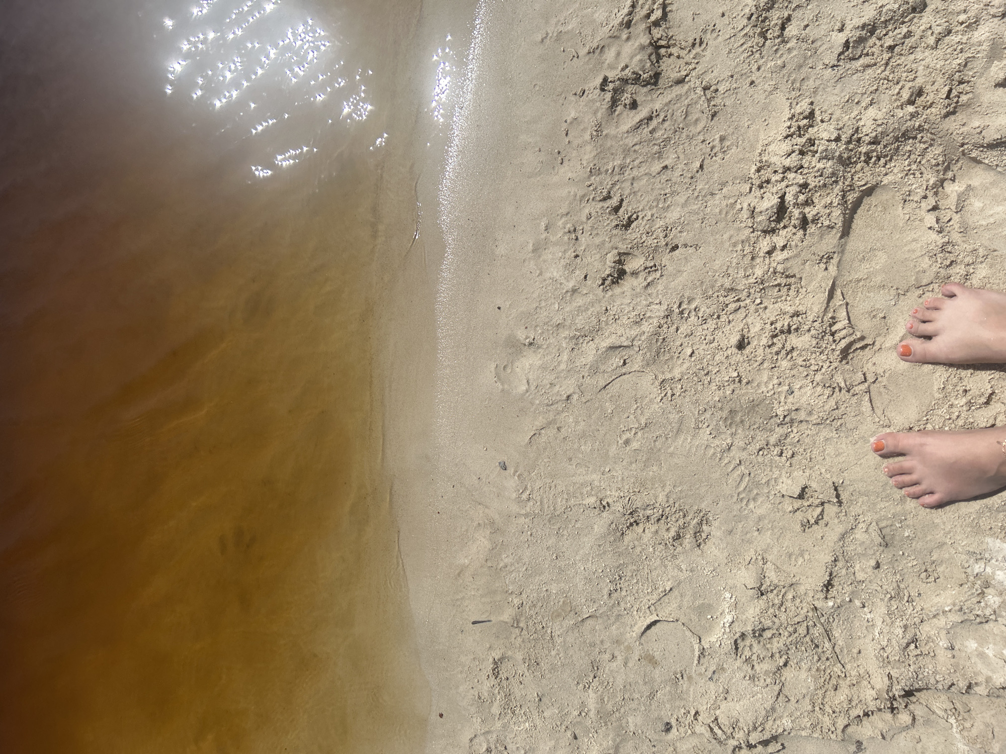 The image shows a sandy beach bordering a body of dark, amber-colored water. The water reflects bright sunlight, creating a speckled, shimmering effect on its surface. The sand is light-colored with visible footprints and some scattered debris. In the lower part of the image, two bare feet with orange nail polish are visible, positioned on the sand near the water's edge. The scene appears to be a natural outdoor environment, likely a beach or shoreline. The location is not specified as Sestroretsk, Russia, and the description is based solely on the visual elements present in the image.