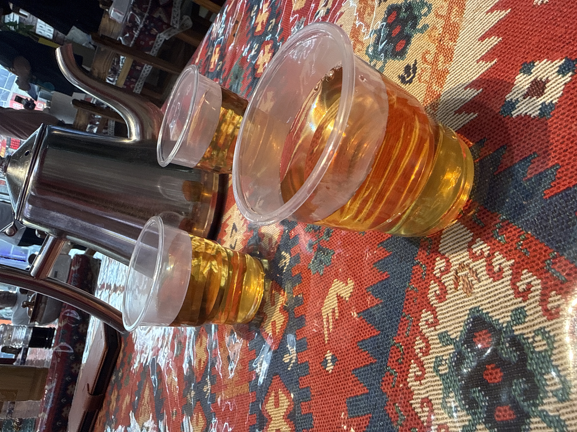 The image depicts three clear plastic cups filled with amber-colored liquid, presumably tea, placed on a table with a red, blue, and beige patterned tablecloth. A large, shiny metal teapot stands behind the cups. In the background, blurred figures of people are visible, along with furniture and other objects, suggesting an indoor setting, possibly a restaurant or cafe. The lighting appears to be ambient, casting subtle reflections on the cups and teapot. The tablecloth has a woven texture with geometric and floral motifs. The scene is captured from a slightly elevated angle, focusing on the drinks and the teapot. There is no specific indication of the location being in Huizhou, China; this detail is not present in the visual information provided.