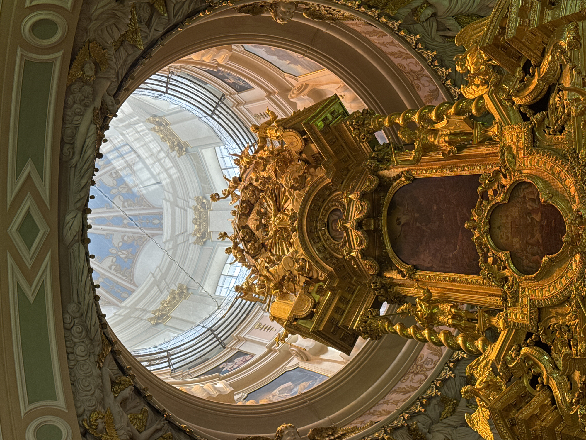 The image shows the opulent interior of a church in Saint Petersburg, Russia, specifically the interior of the St. Isaac's Cathedral's dome. The focal point is a richly gilded altar, adorned with intricate sculptures of angels and other figures. The altar is bathed in natural light streaming from the dome above, which features a stained-glass window and painted murals depicting religious scenes. The surrounding architecture is equally ornate, with elaborate moldings, stucco work, and painted ceilings adding to the grandeur of the space. The overall impression is one of immense wealth and artistic craftsmanship, characteristic of Baroque and Neoclassical styles prevalent in Russian cathedrals.