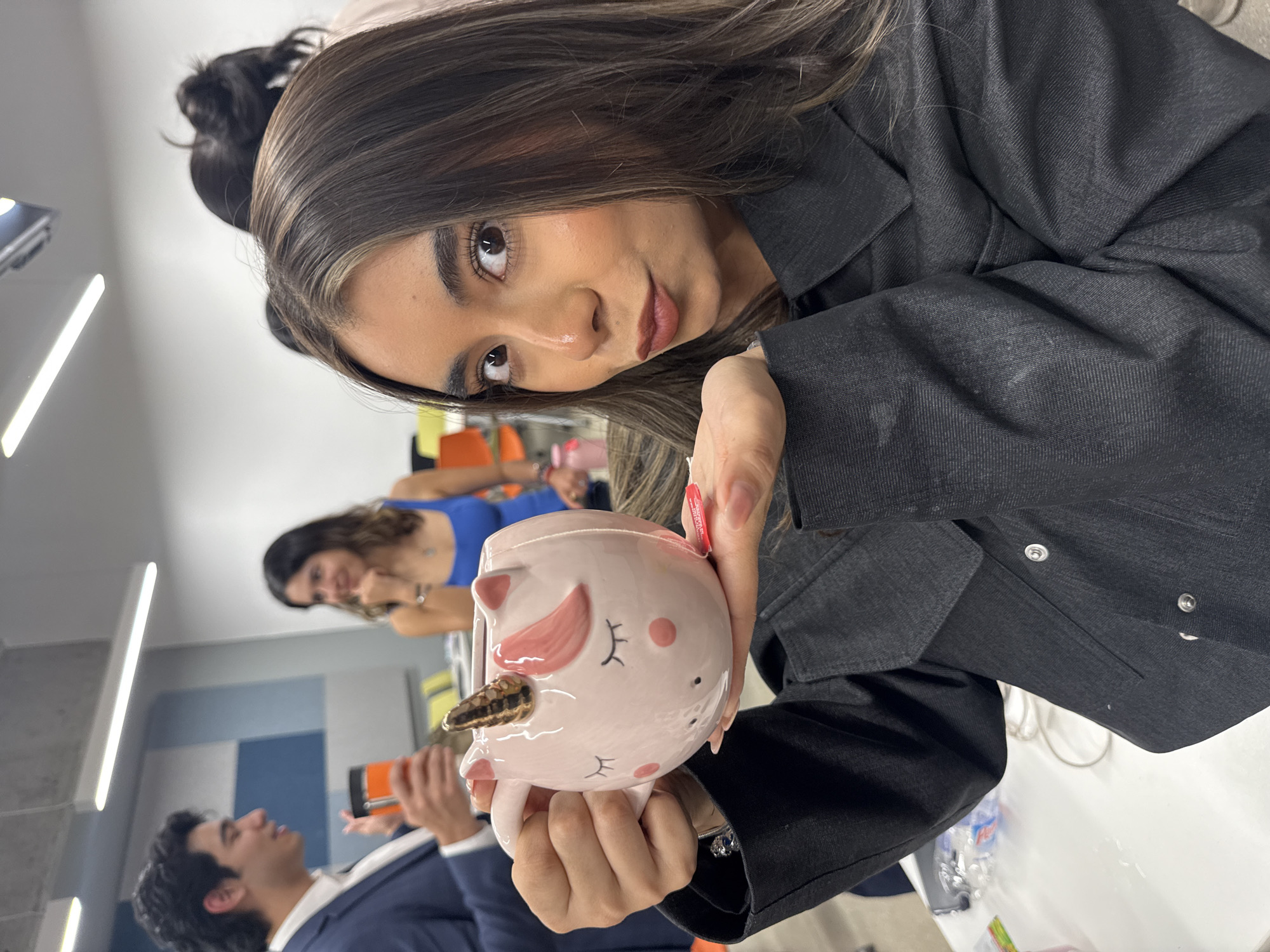 The image, rotated counter-clockwise, depicts an indoor gathering in Mexico City, Mexico. The central subject is a young woman with long, dark brown hair featuring lighter highlights. Her head is tilted, and her dark eyes look directly at the camera with a soft, engaging expression. She has well-defined eyebrows, subtle cheek blush, and soft lips. She is wearing a dark, possibly denim or twill, collared jacket.
She holds a light pink ceramic mug designed like a sleeping unicorn, featuring small pointed ears, rosy cheeks, closed eyes with delicate lashes, and a prominent golden, textured horn. Her fingernails are painted red, and a small, white price tag with red text is attached to the mug's handle, though the text is unreadable.
In the softly blurred background, another woman with dark hair, wearing a blue top, is visible, looking towards the right. Further to the left and behind, a man with dark, curly hair, dressed in a dark suit jacket over a white shirt, holds an orange object and appears to be looking upwards, engaged in conversation.
The setting appears to be a modern indoor space, possibly an office, classroom, or event hall, characterized by white tables, light-colored walls with blue and gray sections, and linear ceiling lighting. Several water bottles and white cables are sparsely visible on the tables. The bright, even lighting suggests it is daytime. The atmosphere seems relaxed and social.