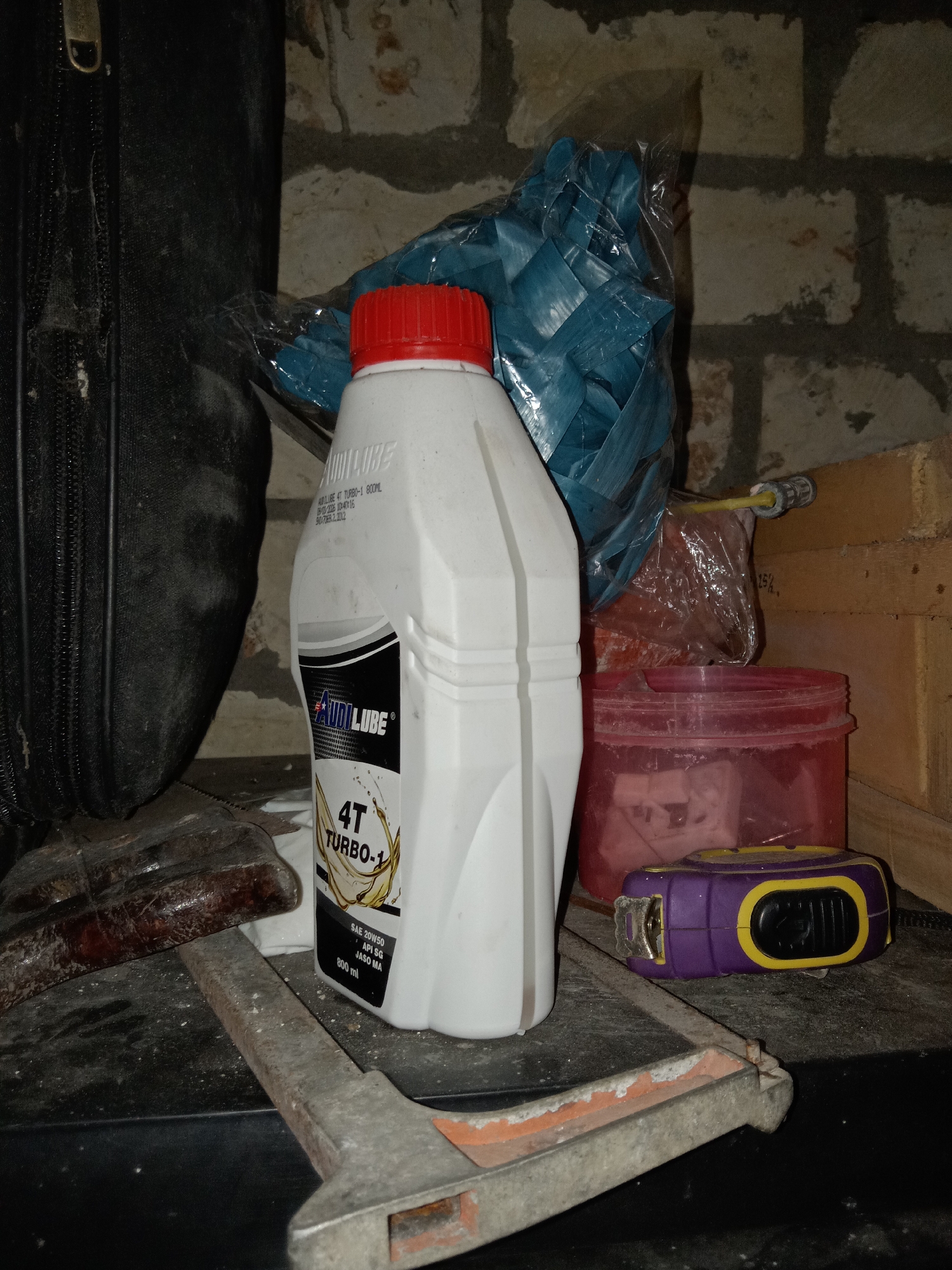 An indoor, low-light scene depicts a collection of miscellaneous items on a dark, possibly concrete or stone surface. In the foreground, a metal tool, possibly a wrench or clamp, with orange paint remnants, lies horizontally. To its right, a purple and yellow measuring tape rests on the surface. Further back and to the left, a black, zippered case or bag is partially visible. Dominating the center of the image is a white plastic bottle with a red cap, containing what appears to be oil or lubricant, with a label indicating "AUDILUBE 4T TURBO-1 800 ML". Behind the bottle, a blue woven plastic bag is visible, partially covered by clear plastic. To the right of the oil bottle, a transparent red plastic container is present. The background consists of a brick wall, illuminated by a low-light source. The scene appears to be a storage area, potentially a workshop or garage, within a building. There is no indication of a specific location in Jumput, Indonesia, or any discernible human presence or activity.