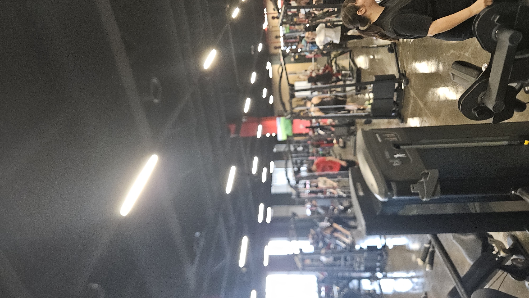 The image depicts a bustling indoor gym located in Plovdiv, Bulgaria. The perspective looks across a wide exercise area, showcasing numerous fitness machines and individuals engaged in physical activity under bright artificial lighting.
In the mid-ground on the right, a woman with dark hair, wearing a black short-sleeved top, is visible, appearing to use a leg press or similar strength training machine, with black weight plates partially visible. Adjacent to her and further into the foreground is the side profile of a black treadmill with a grey control panel. The floor beneath the equipment and people is a light, somewhat reflective brown or beige.
The background extends into a large, well-equipped space filled with rows of various gym apparatus. Other gym-goers can be seen utilizing different cardio and strength machines, though their faces are indistinct. Some equipment has red elements.
The ceiling, which takes up a significant portion of the left side of the frame, is dark grey or black and features numerous bright, rectangular LED light fixtures arranged in parallel rows, providing strong, even illumination throughout the facility. These lights create noticeable lens flares and reflections.
The overall scene suggests a busy, modern fitness center during an active period. No specific text or legible branding is visible on any of the equipment or within the gym environment.