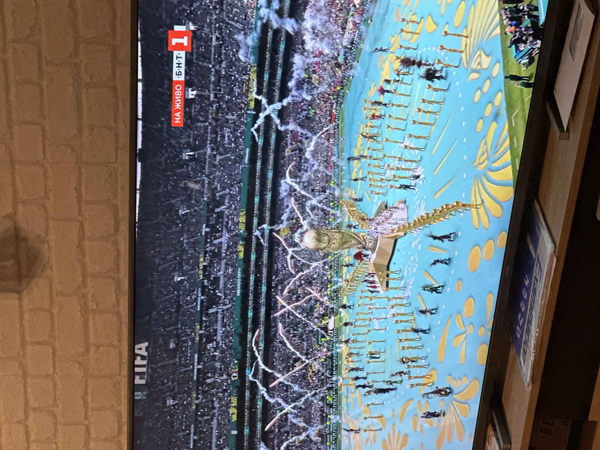 A television screen displays a live broadcast of an event, likely a sporting ceremony. The broadcast features a stadium filled with spectators, with a large open area in the foreground. This area is decorated with a bright blue surface and yellow floral patterns. Numerous individuals, dressed in gold costumes and some in dark, tribal-inspired outfits, are positioned in rows and scattered across the blue area, appearing to perform or be part of a presentation. A large, golden trophy, resembling the FIFA World Cup, stands prominently on a raised platform in the center. Fireworks and smoke effects are visible in the background, originating from the stadium and above the performers. Overlaying the broadcast are graphical elements: "MEXICO CITY" is repeatedly visible on the stadium advertising boards, and in the upper right corner, a red banner displays "НА ЖИВО Б.Н.Т." (meaning "LIVE B.N.T.") next to a red square with the number "1" inside, indicating a live broadcast on the BNT channel. The television itself is placed on a wooden stand, with a brick-patterned wall visible behind it.