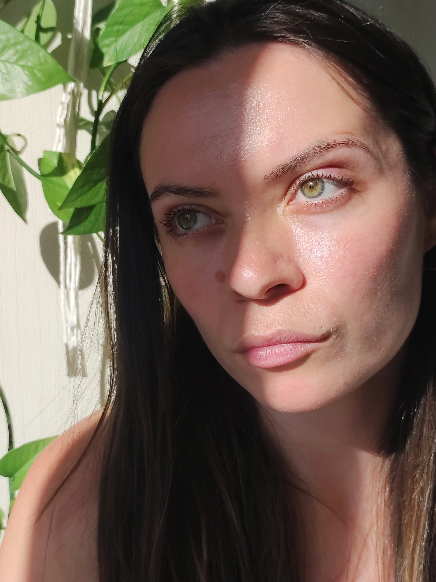 A close-up shot of a woman with long, dark brown hair and light green eyes. A shadow crosses her forehead and cheek, cast by an unseen source. She has fair skin and a mole near her nose. The subject appears to be partially lit by natural light. To the left of her face are green leaves of a potted plant. The background is an off-white wall. The location cannot be determined from this image.