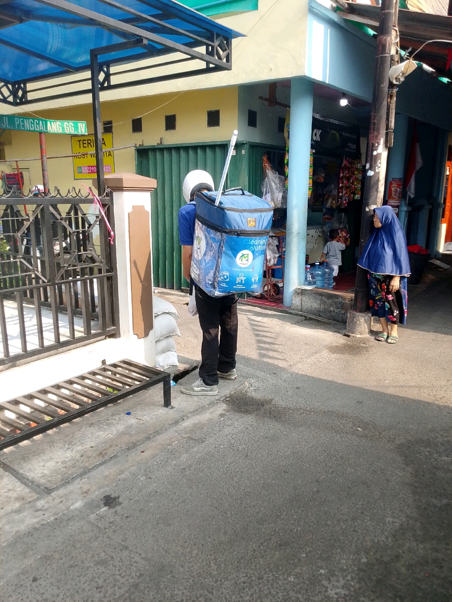 The image captures a street scene in East Jakarta, Indonesia, specifically on Jalan Penggalang GG IV. A delivery person, wearing a blue shirt and black pants with a white helmet, is seen from behind carrying a large blue insulated delivery bag on their back. The bag has graphics and text, including "Cleaning Solution." To the left of the delivery person is a black metal fence.
In the background, a yellow building is visible with a green metal gate. A sign on the building reads "TERIMA KOST WITA" along with a phone number. A blue awning extends over a portion of the street.
To the right of the delivery person, a woman wearing a blue hijab and a floral patterned skirt is standing next to a utility pole. Further in the background, a small child in a grey shirt is near some large blue water bottles inside a shop with a blue facade. The ground is paved with asphalt and concrete. The lighting suggests it is daytime, with shadows cast by the sun.