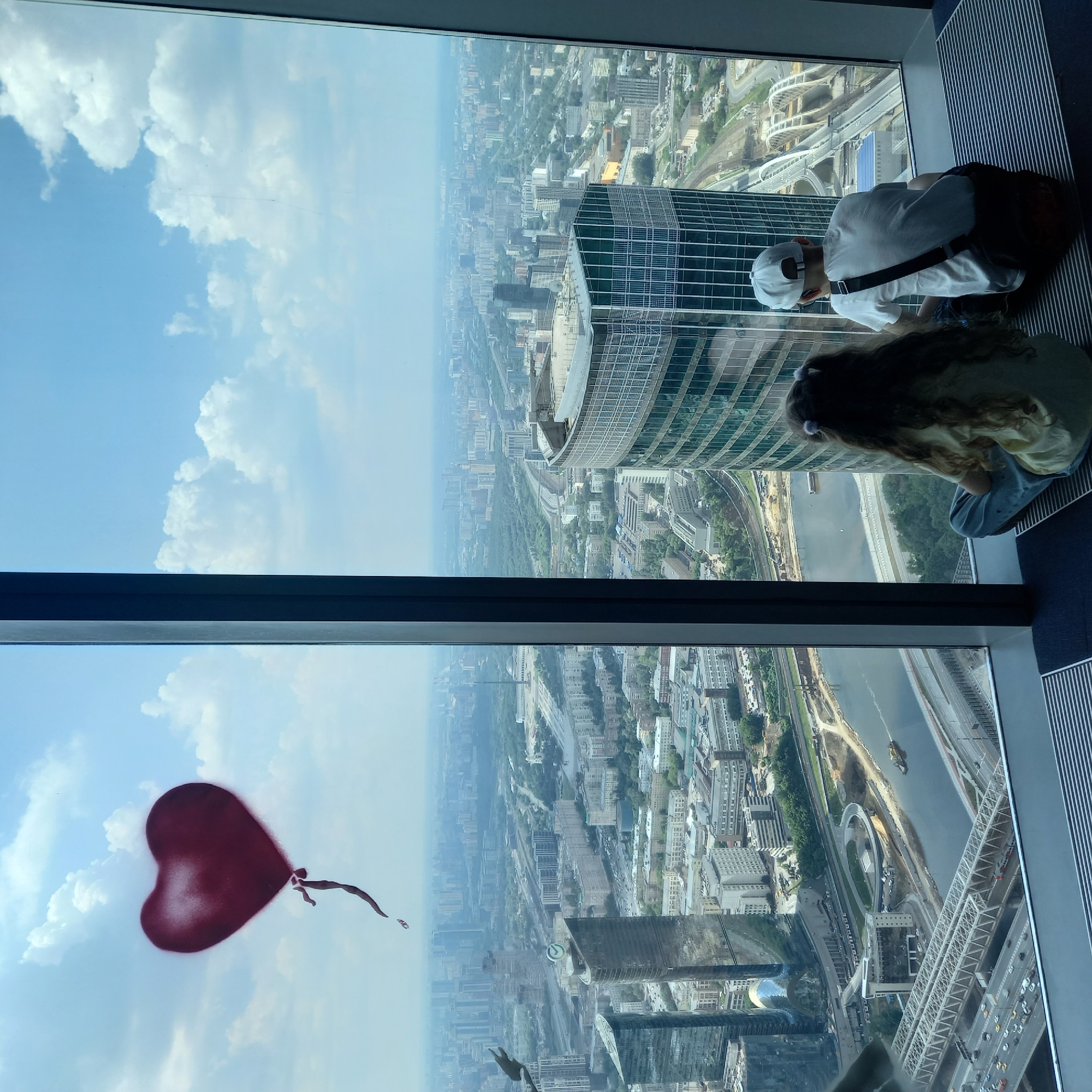 The image captures a panoramic view of a sprawling cityscape, viewed from a high vantage point through a large window. In the foreground, two children are seated on the floor, their backs to the camera, gazing out at the urban landscape. The child on the left has long, dark, wavy hair with two small purple hair ties, and is wearing a light-colored shirt and blue pants. The child on the right wears a white t-shirt and a white baseball cap, with a dark crossbody bag slung over their shoulder.
The city below is densely packed with buildings of varying architectural styles and heights, interspersed with green spaces and roadways. A prominent river winds through the city, crossed by several bridges. In the sky, a large, red, heart-shaped balloon with a trailing string is depicted, appearing as if it's floating in the distance, though it is likely a superimposed graphic element. The sky is partly cloudy with a clear blue expanse. The overall impression is one of awe at the vastness of the city, with the children appearing contemplative as they observe the scene.