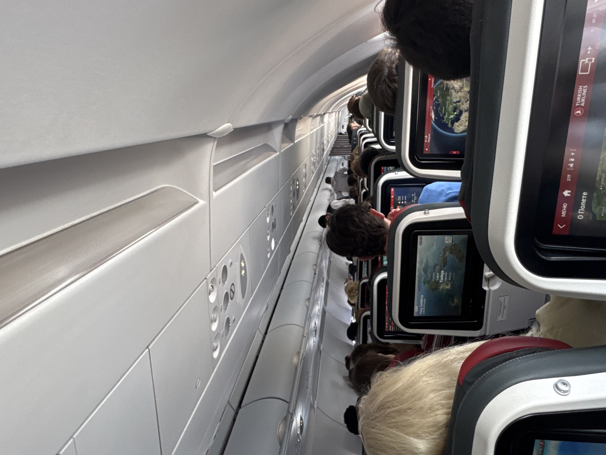 The image shows the interior of an airplane cabin during a flight. Rows of seats are visible, occupied by passengers. Each seat appears to have an individual screen, some displaying information about Turkish Airlines. The interior is brightly lit, and the overhead compartments are visible. The screens display details such as the flight path, Turkish Airlines logo, and other flight information. The text on the screens is in multiple languages. The location is not in Saint Petersburg, Russia. The screens of the plane indicate that it is probably a Turkish Airlines flight.